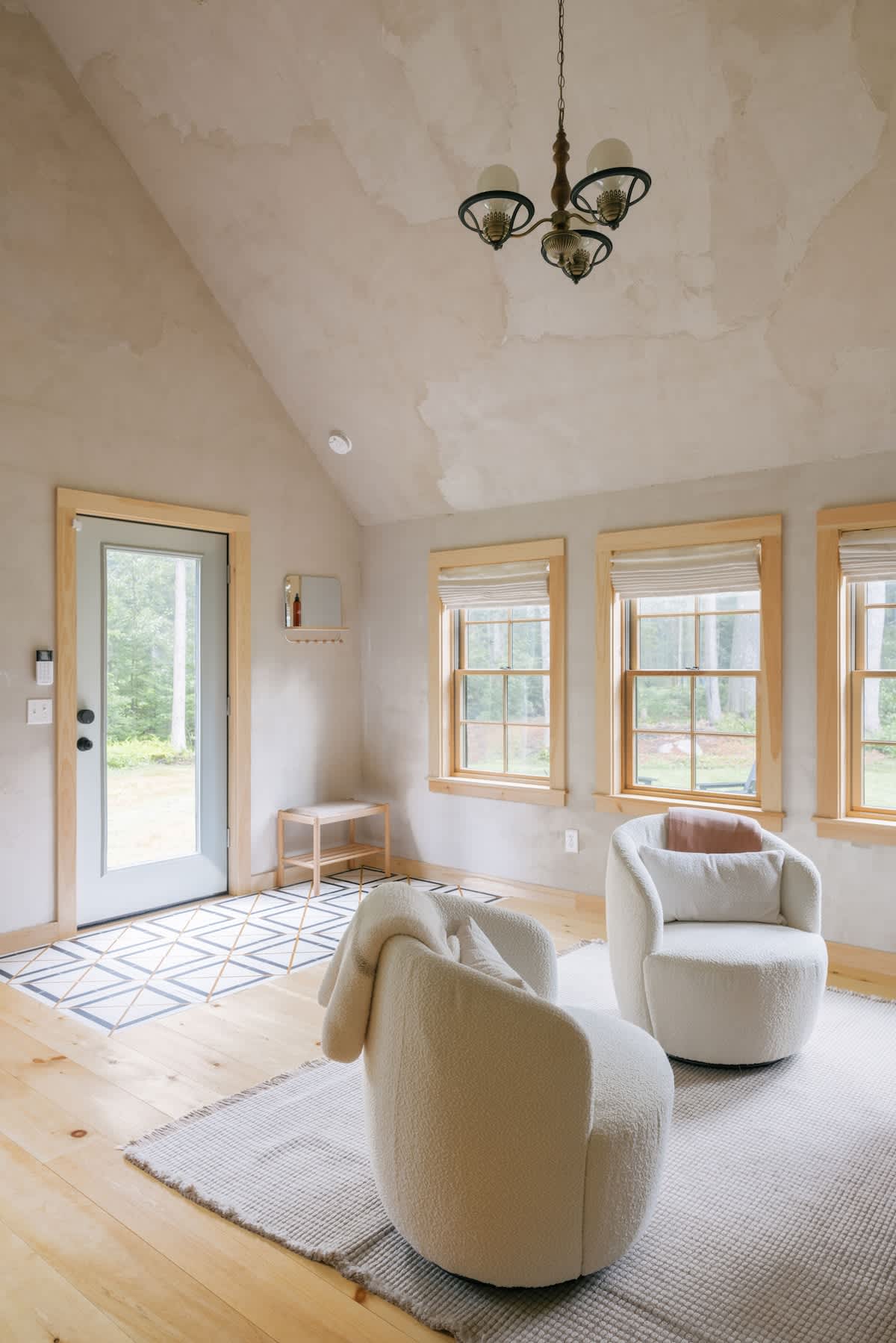 A bright, minimalist living space with a vaulted ceiling and light wood floors, featuring two plush white swivel armchairs on a textured rug. Large windows with wood trim offer views of the surrounding trees, and a light blue door provides outdoor access.
