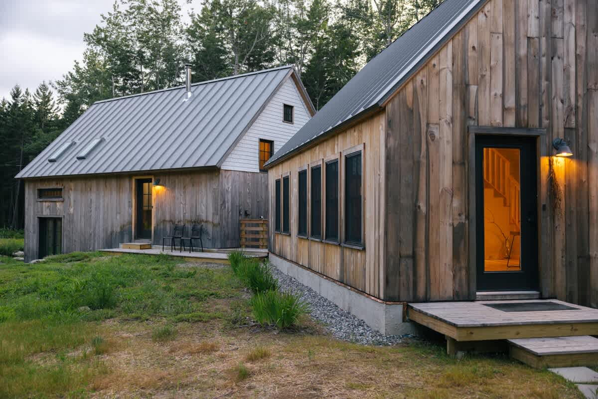 Two rustic wooden cabins with gray metal roofs stand amidst a grassy yard and trees. The right cabin has a small wooden deck and a lit doorway, while the left has a larger deck with chairs.