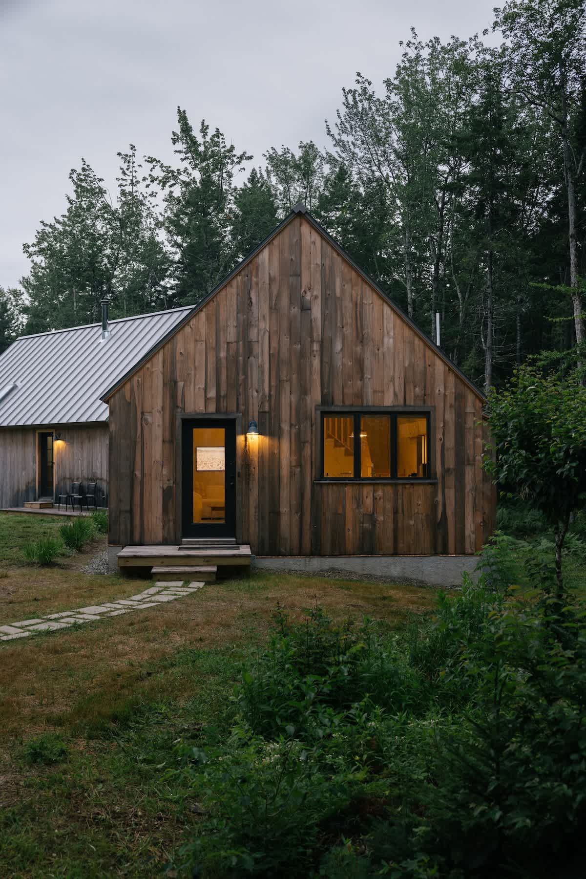 A rustic wooden cabin with a gray metal roof and warm interior lights stands at dusk, with a stone path leading to its entrance. Dark trees frame the background.