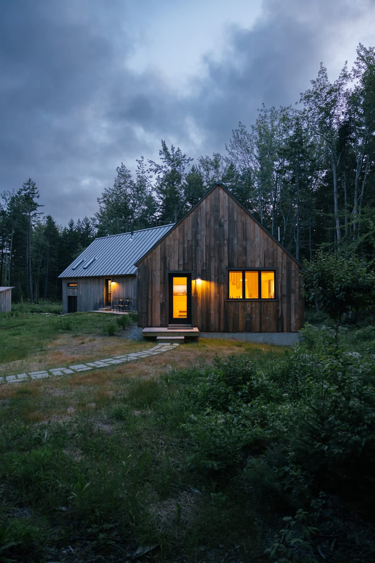 A rustic wooden cabin with a gray metal roof and warm interior lights glows at dusk, surrounded by dark trees and a grassy path. A small wooden deck leads to the entrance.