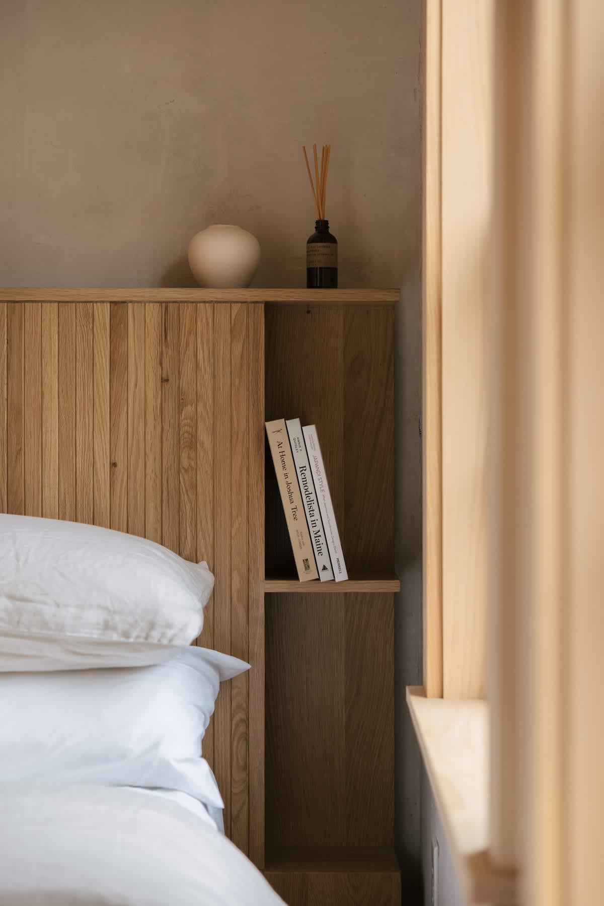 Detail shot of the bedroom's slatted wooden headboard with integrated shelving, displaying books, a minimalist vase, and a reed diffuser, next to a window with natural light.