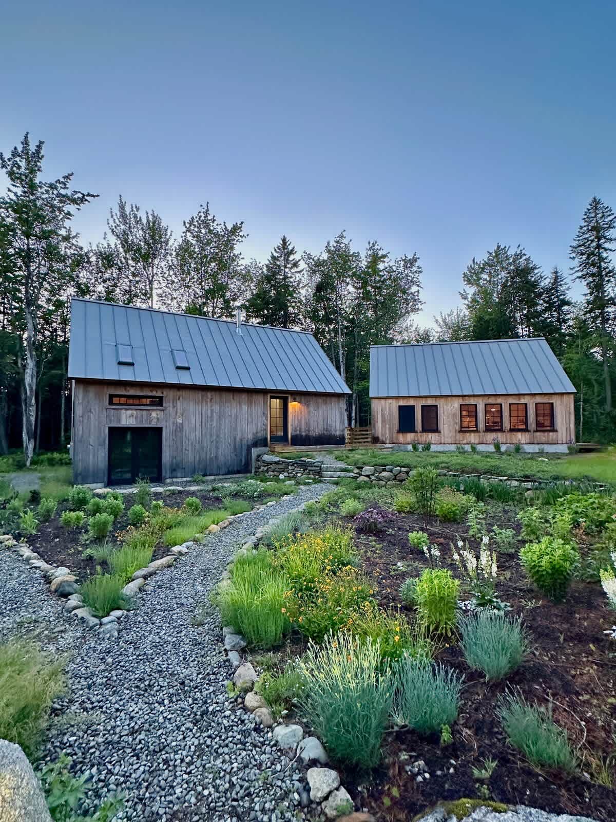 Two rustic wooden cabins with gray metal roofs overlook a landscaped garden with a gravel path and various plants. The sky is a soft twilight blue.