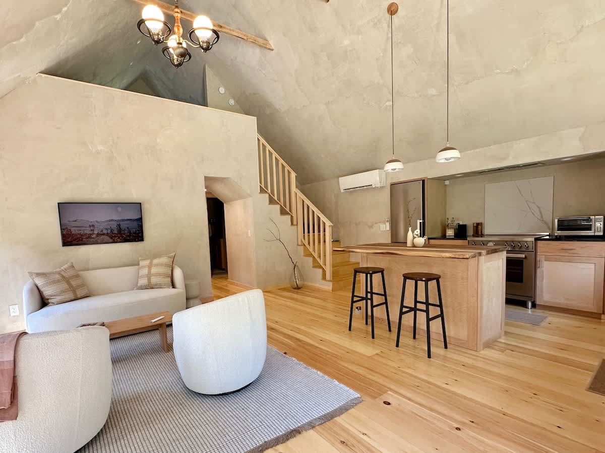 An open-concept living space with a vaulted ceiling and light textured walls, featuring a cream sofa, two round white armchairs, a wooden coffee table, and a view into the kitchen with a wooden island and bar stools. A wooden staircase leads to an upper level.