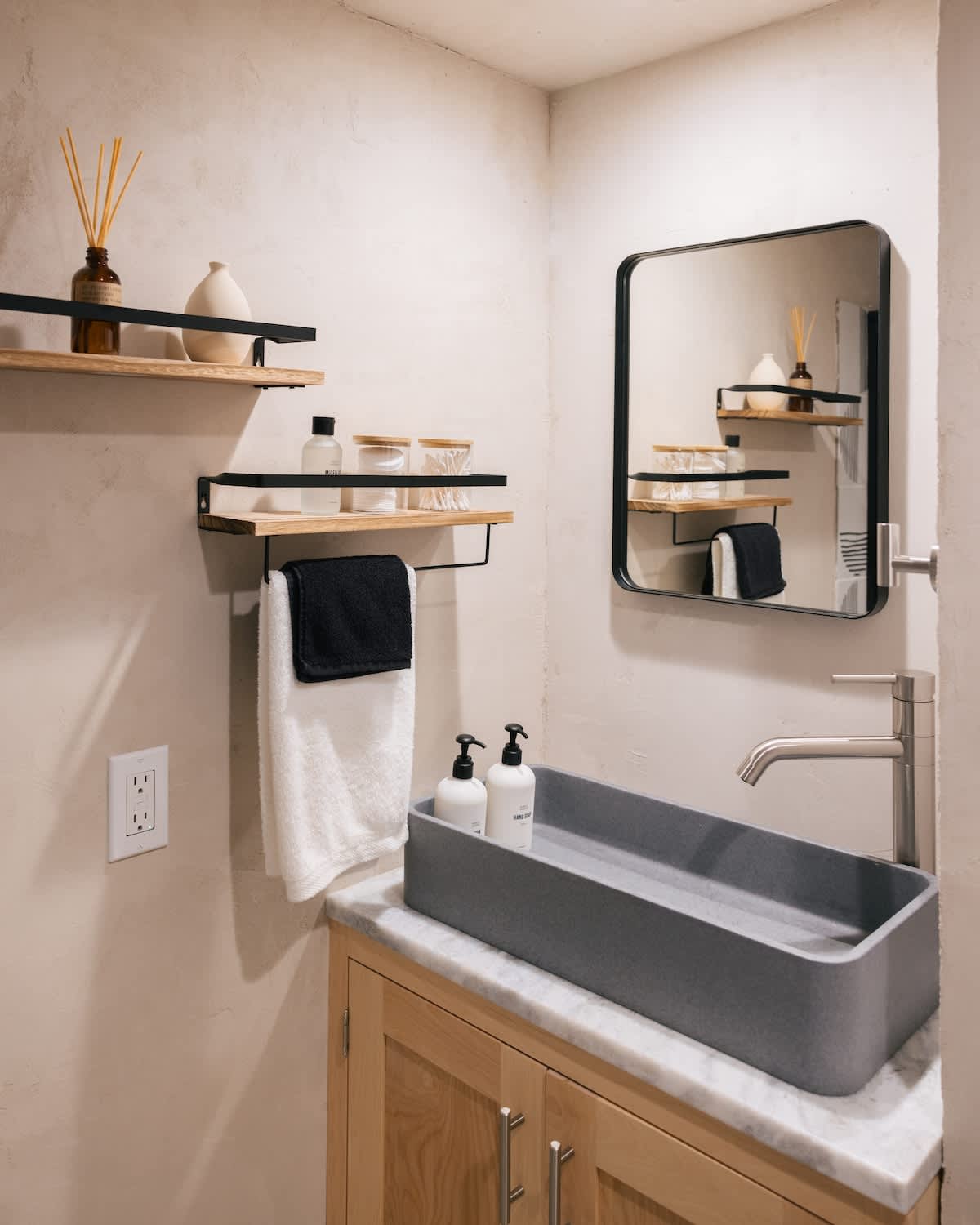 A contemporary bathroom vanity features a unique gray trough sink set on a marble countertop, a light wooden cabinet, a rectangular black-framed mirror, and two floating shelves with decor and toiletries.