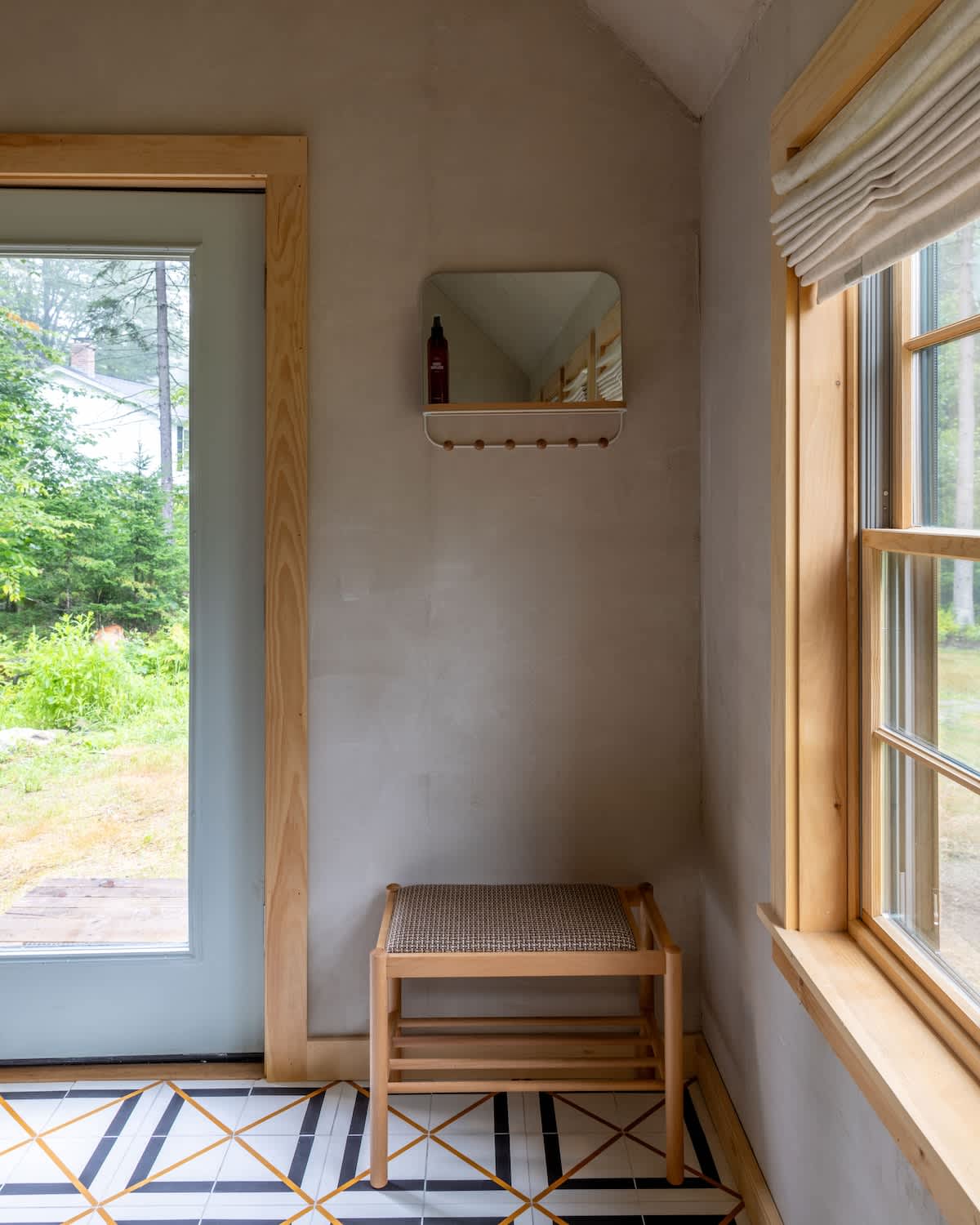 A bright entryway features a patterned tile floor, a wooden bench, and a square mirror with hooks on a light gray wall. A large window and a light blue door offer outdoor views.