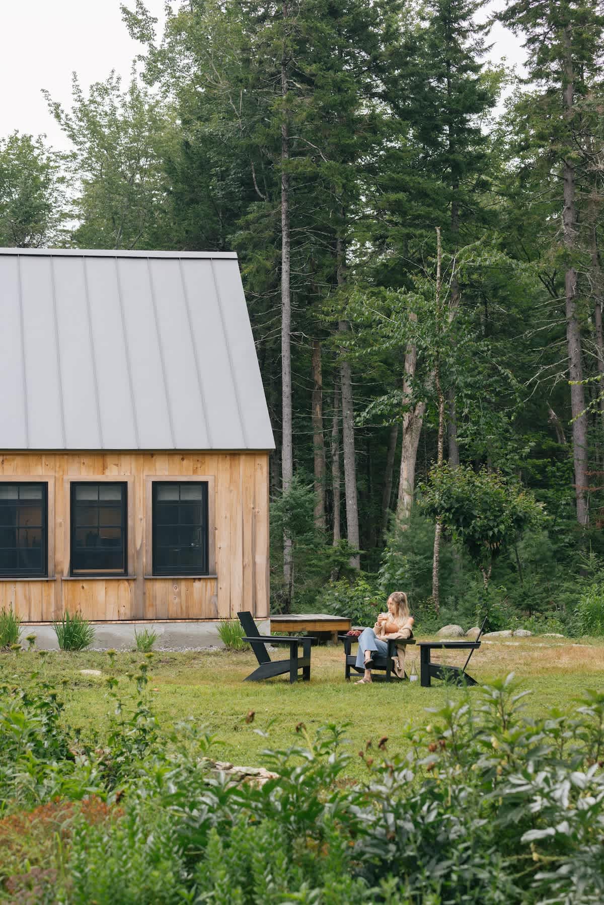 A woman sits in a black Adirondack chair in a grassy yard, facing a rustic wooden cabin with a gray metal roof and black-framed windows. Tall trees line the background.