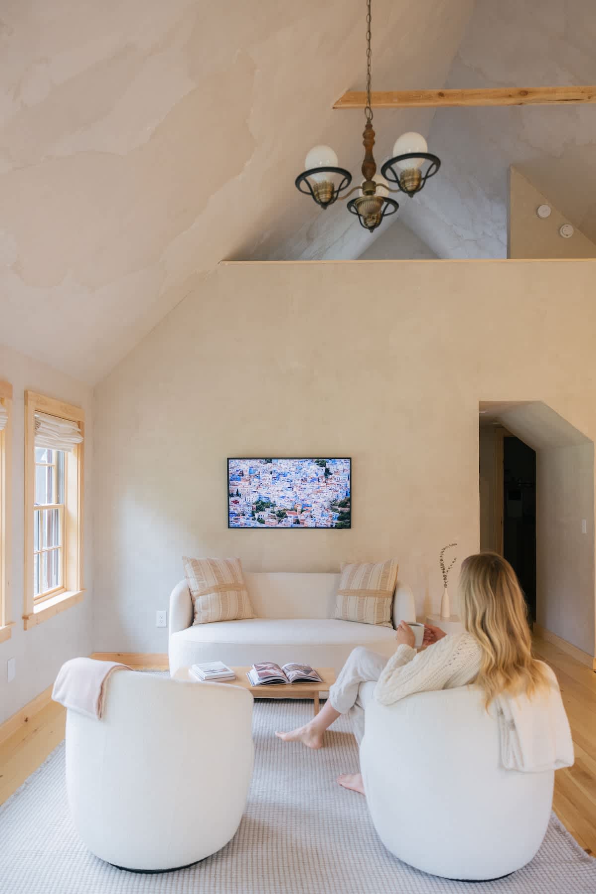 A cozy living room with a vaulted ceiling and light textured walls, featuring a cream sofa with striped pillows, two round white armchairs, a wooden coffee table, and a wall-mounted TV displaying a colorful image.