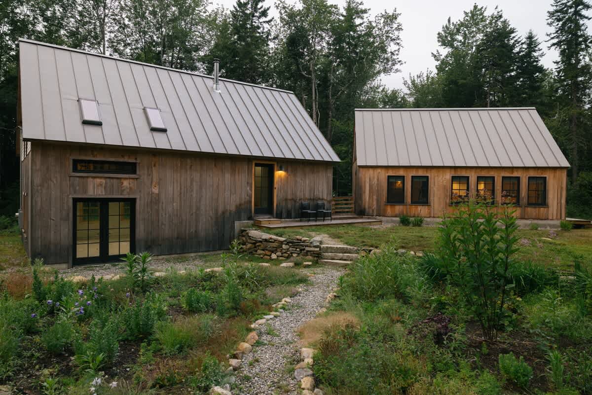 An aerial view showcases two modern rustic cabins with vertical wooden siding and gray metal roofs, connected by a winding gravel path through a wild, grassy landscape with surrounding trees.