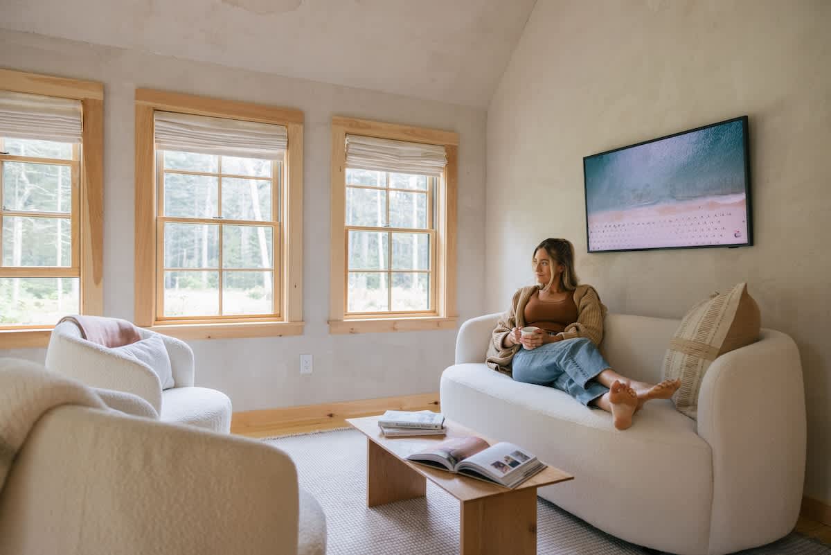 A bright living room with a woman relaxing on a cream sofa, facing two large windows with wooden frames and blinds that offer a view of lush greenery outside. The room features light hardwood floors and a wooden coffee table.