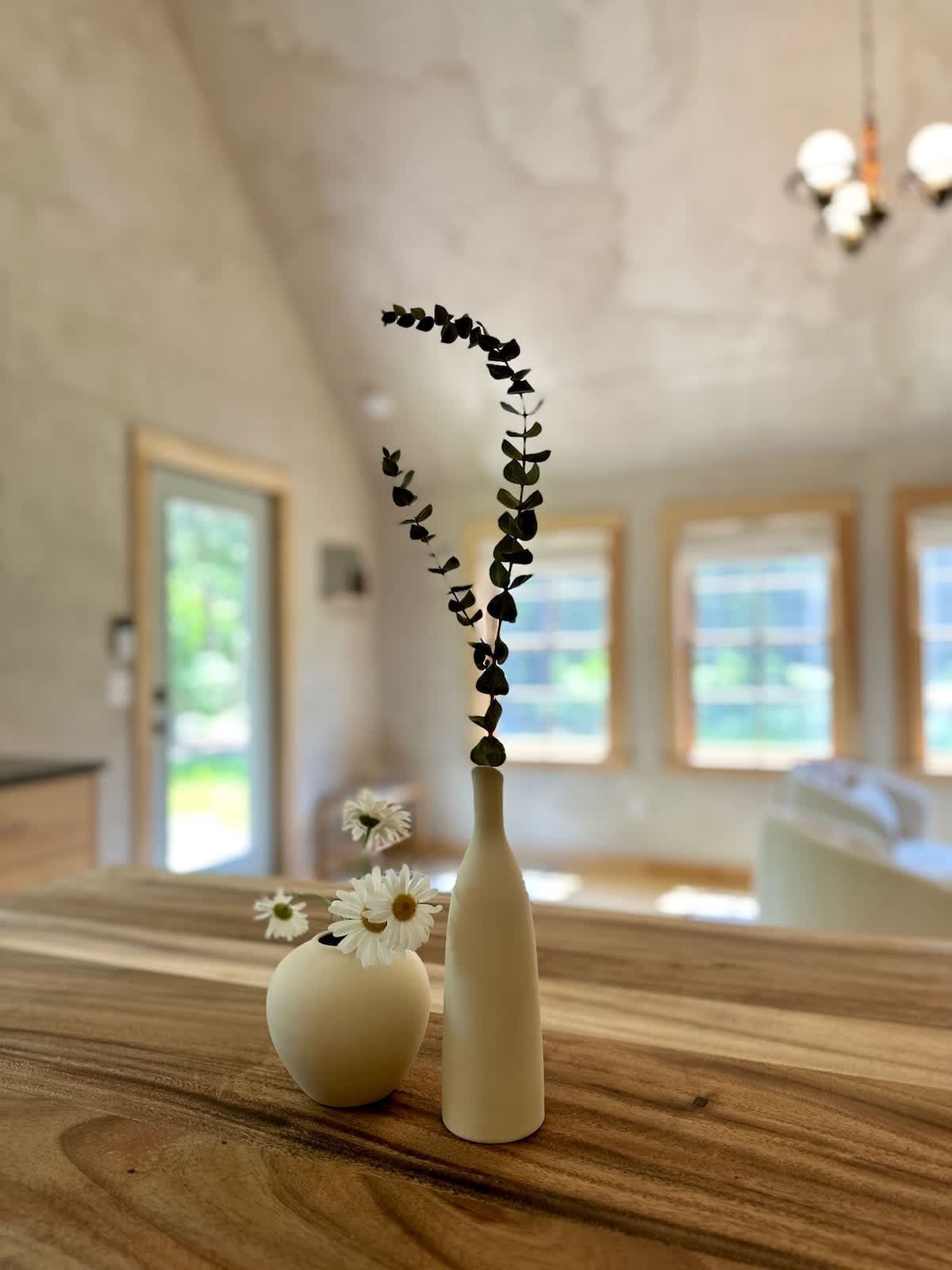 A close-up shot highlights two minimalist ceramic vases, one holding eucalyptus and the other daisies, placed on a warm wooden table, with a blurred background of a bright interior space.