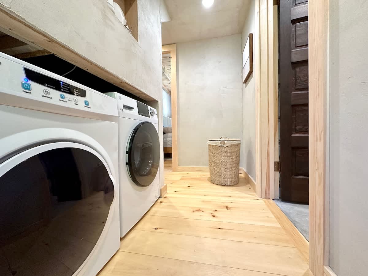A narrow laundry area featuring a white front-loading washer and dryer stacked side-by-side under a wooden shelf, with light wooden floors and a woven laundry basket visible further down the hall.