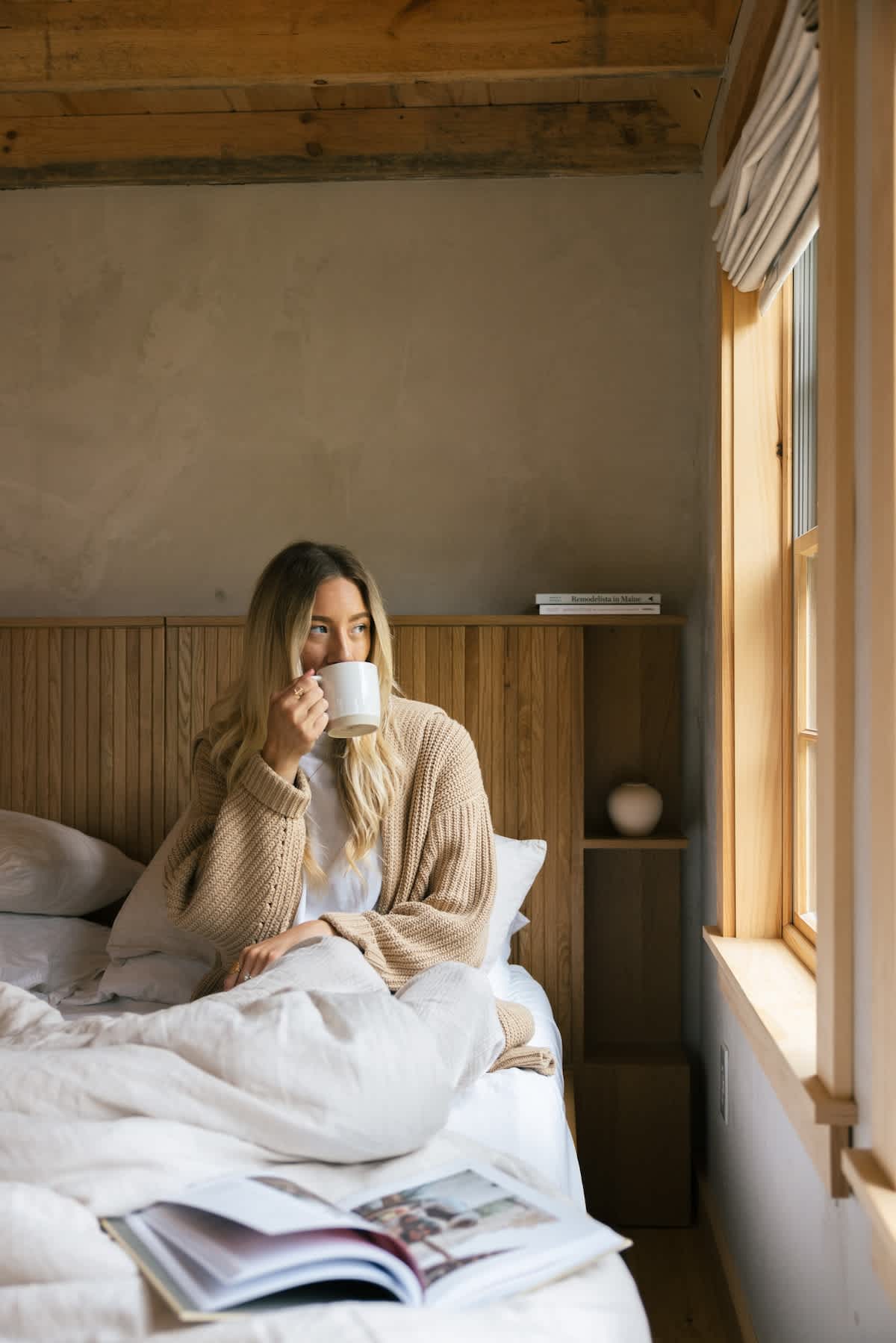 Bright bedroom with exposed wooden ceiling beams and light-colored walls, featuring a bed with a slatted wooden headboard and a person enjoying a drink while looking out the window.
