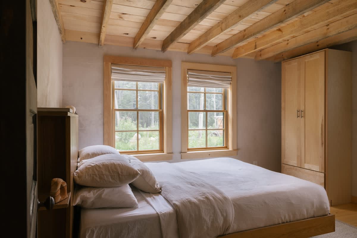 A cozy bedroom features a queen-sized bed with white linens, two windows with natural wood frames and blinds, exposed wooden ceiling beams, and a light wood wardrobe, creating a rustic yet bright ambiance.
