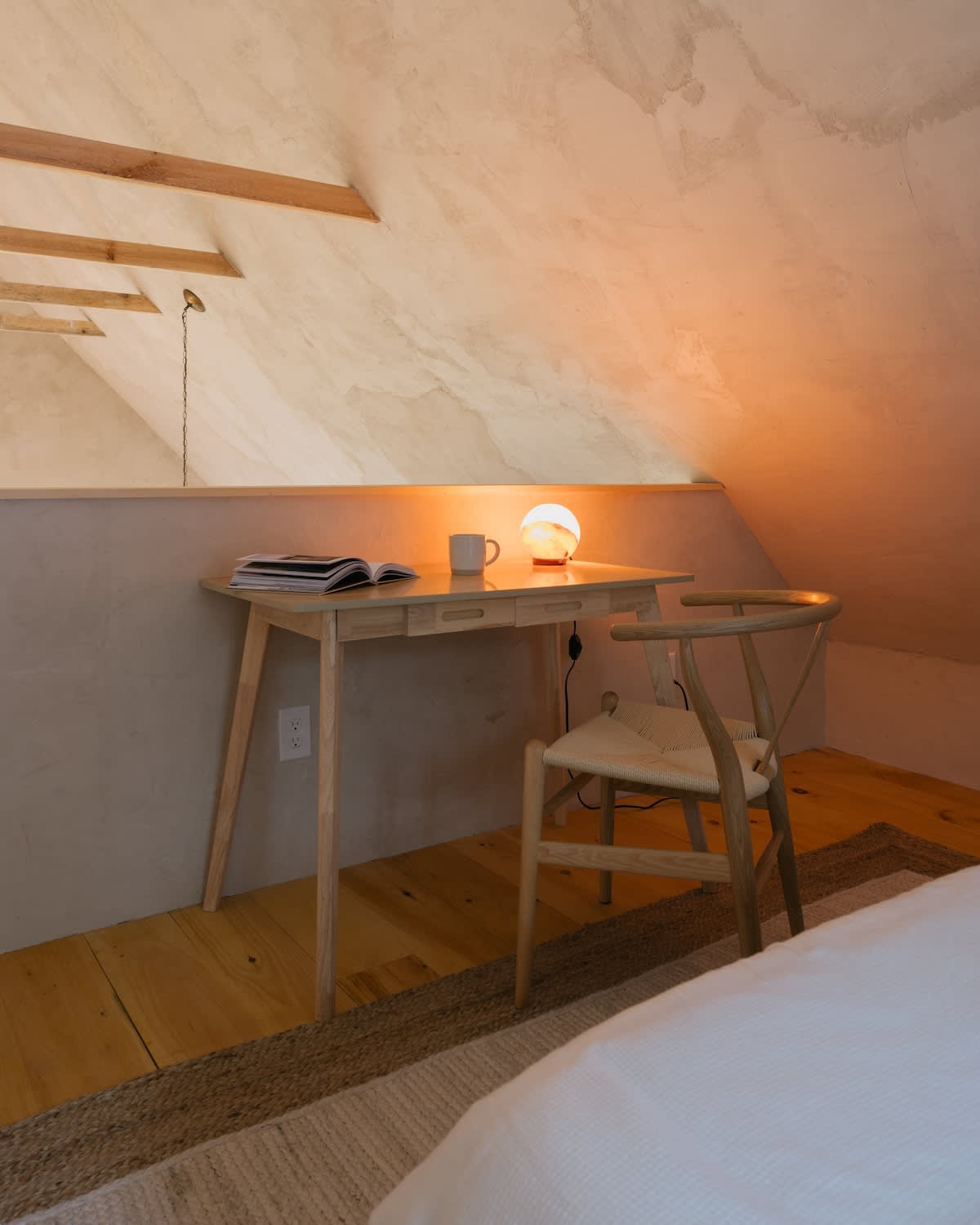 A cozy workspace features a light wooden desk with a small lamp and books, paired with a classic wooden wishbone chair, set against a textured wall with a sloped ceiling, ideal for remote work.