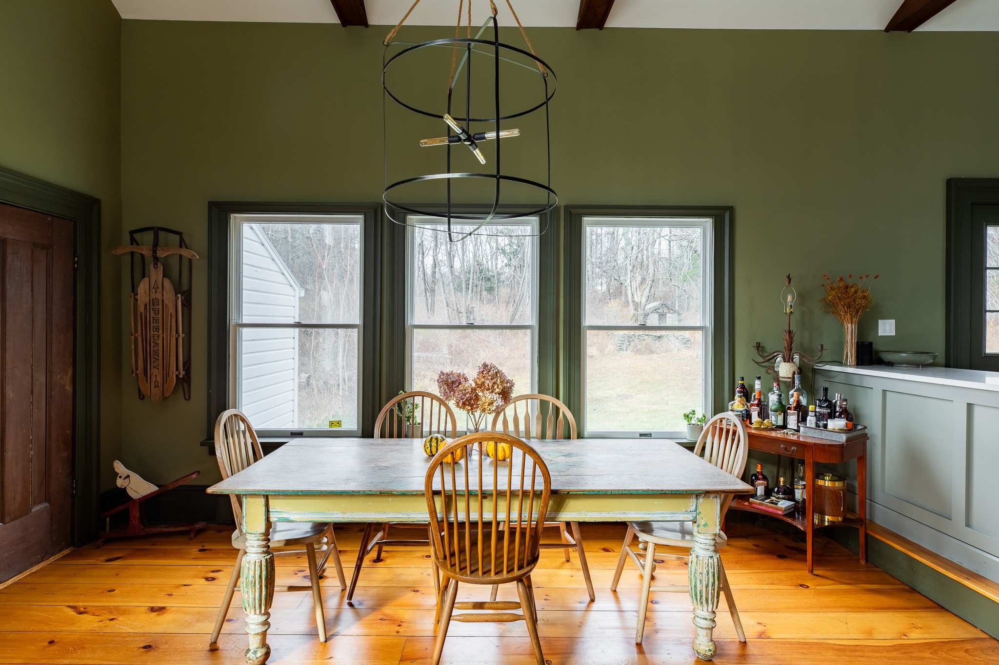 A rustic dining area with olive green walls and exposed beams features a distressed wooden table, spindle-back chairs, and large windows overlooking a grassy backyard.
