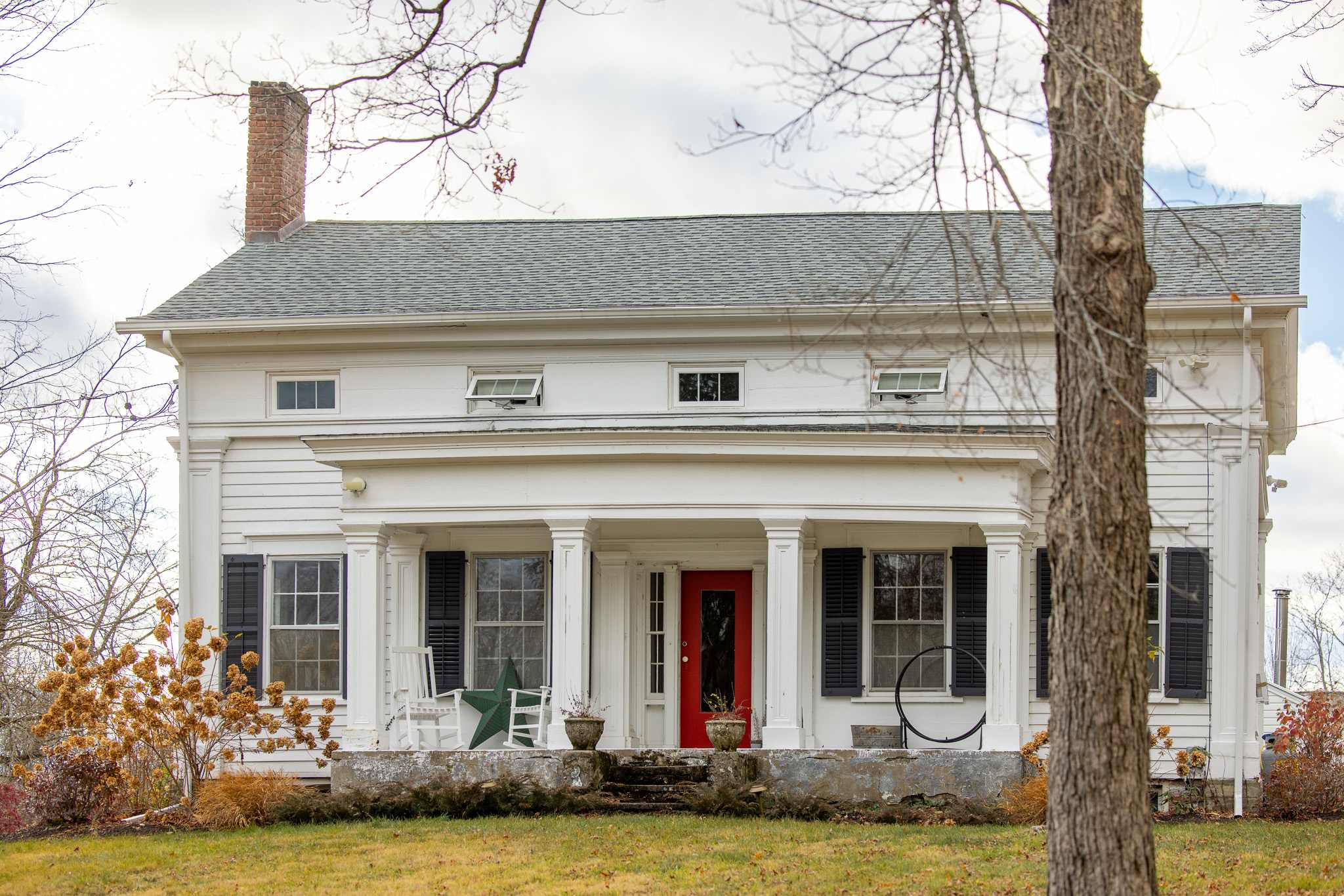 The welcoming front facade of a traditional white farmhouse, featuring a vibrant red front door, a covered porch with classic white columns, and black window shutters, set against a backdrop of bare trees.