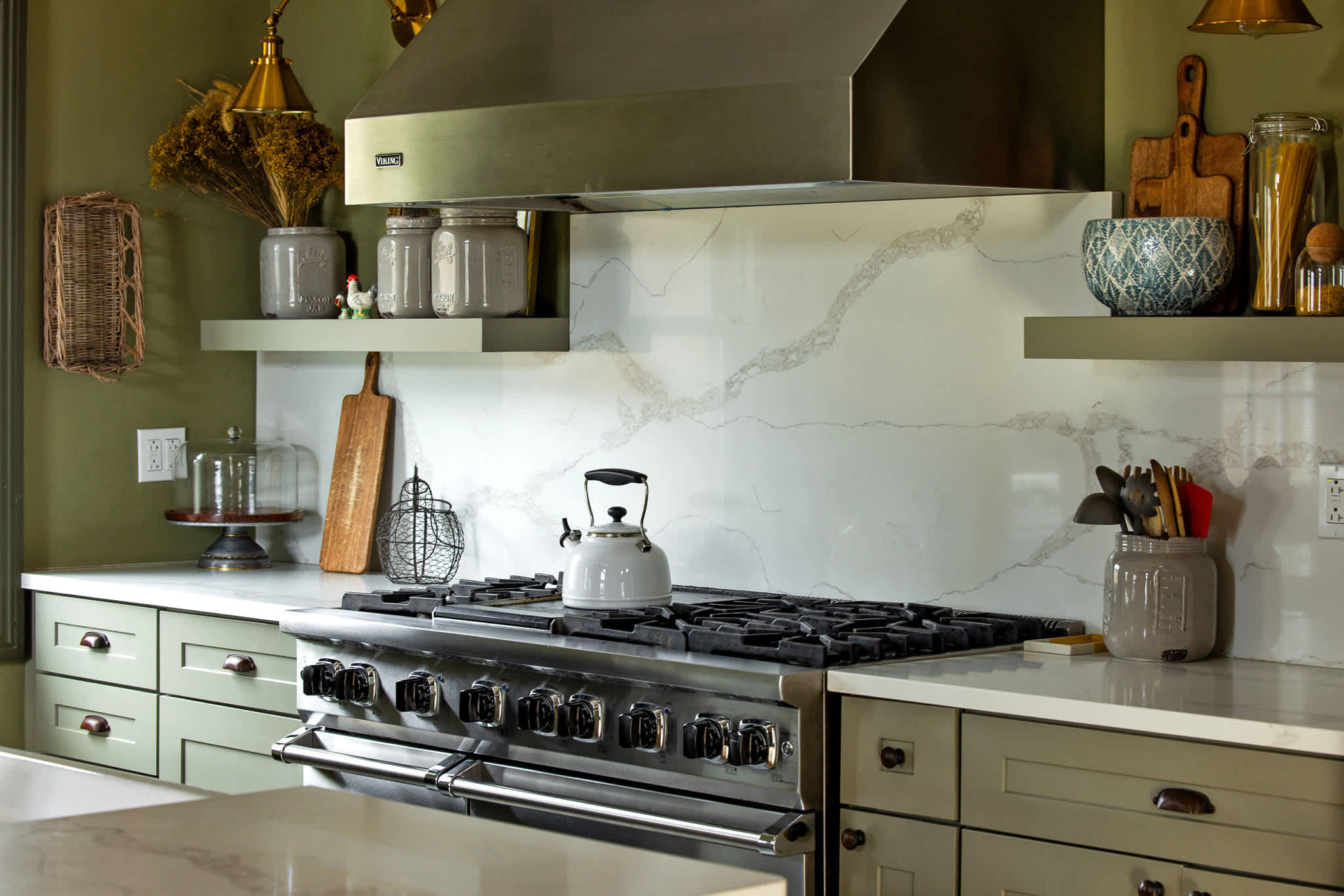 A detailed shot of the kitchen's cooking area highlights a professional-grade stainless steel range with a matching hood, a white kettle, and open shelving against a white marble-patterned backsplash.