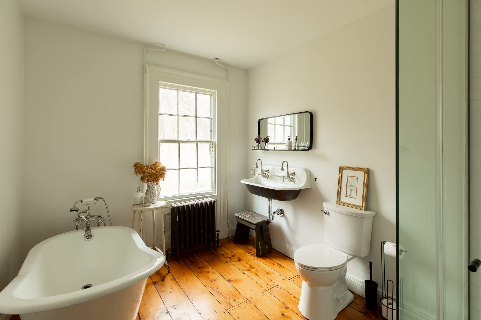 A spacious bathroom with light walls and warm hardwood floors, featuring a white clawfoot tub, a wall-mounted double sink with a dark base, and a toilet. A large window with a radiator below provides natural light.
