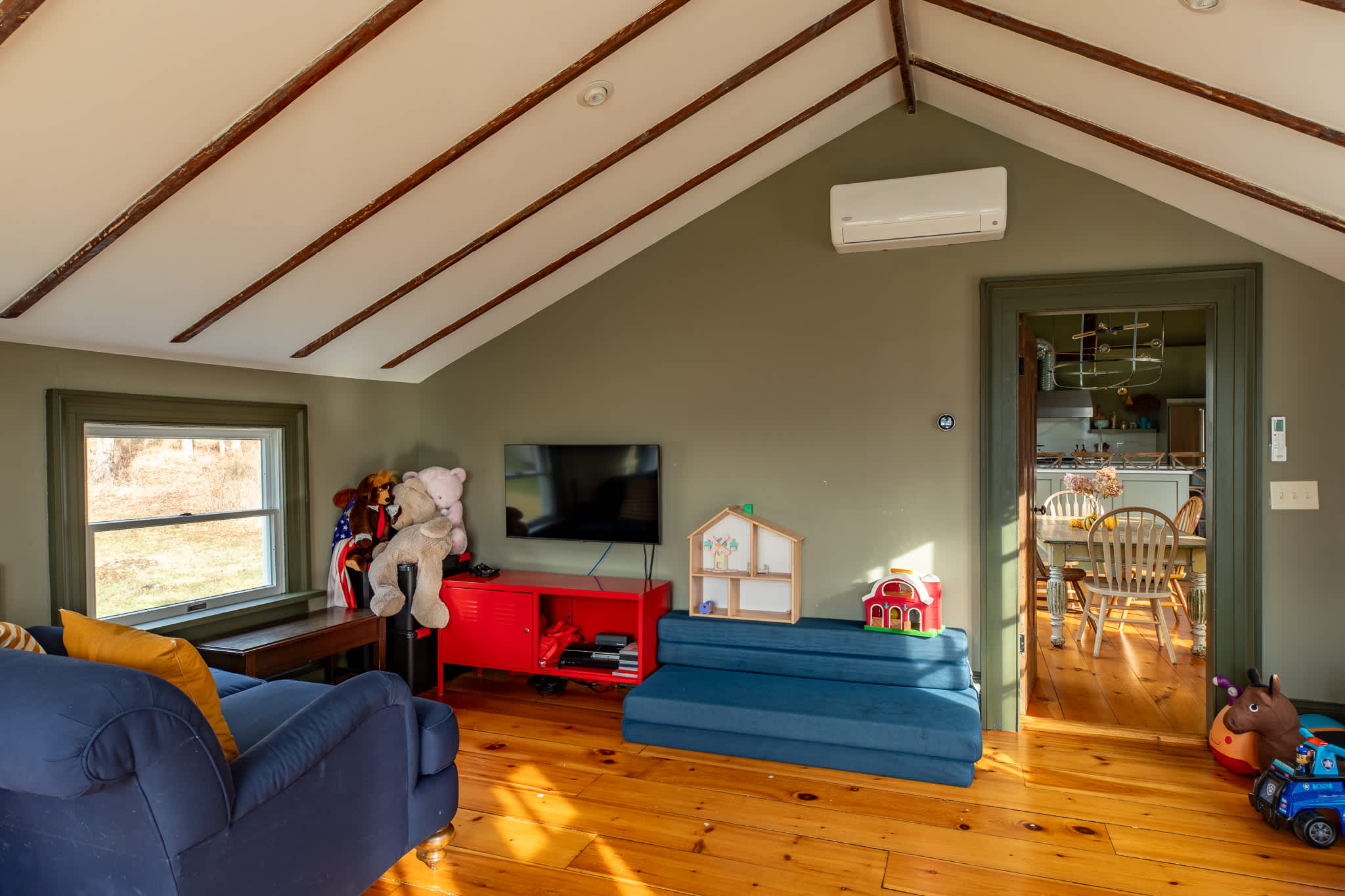 A cozy playroom with an angled ceiling and exposed beams features a blue sofa, a red media console with a TV, and various children's toys, with a glimpse of the kitchen through an open doorway.
