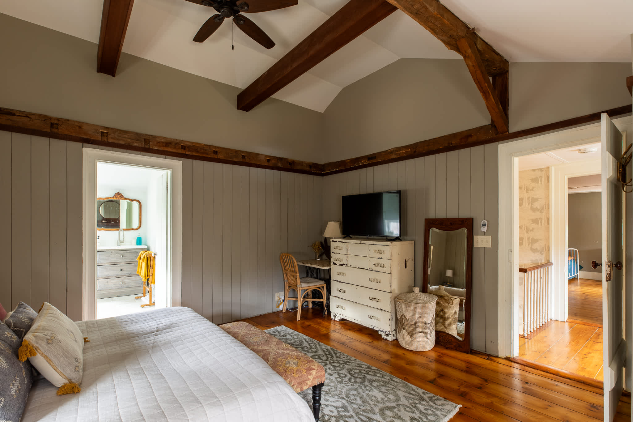 A rustic bedroom with sloped ceilings, exposed wooden beams, and light green paneled walls, featuring a bed, a white distressed dresser with a TV, and a full-length mirror. An open doorway leads to an en-suite bathroom.