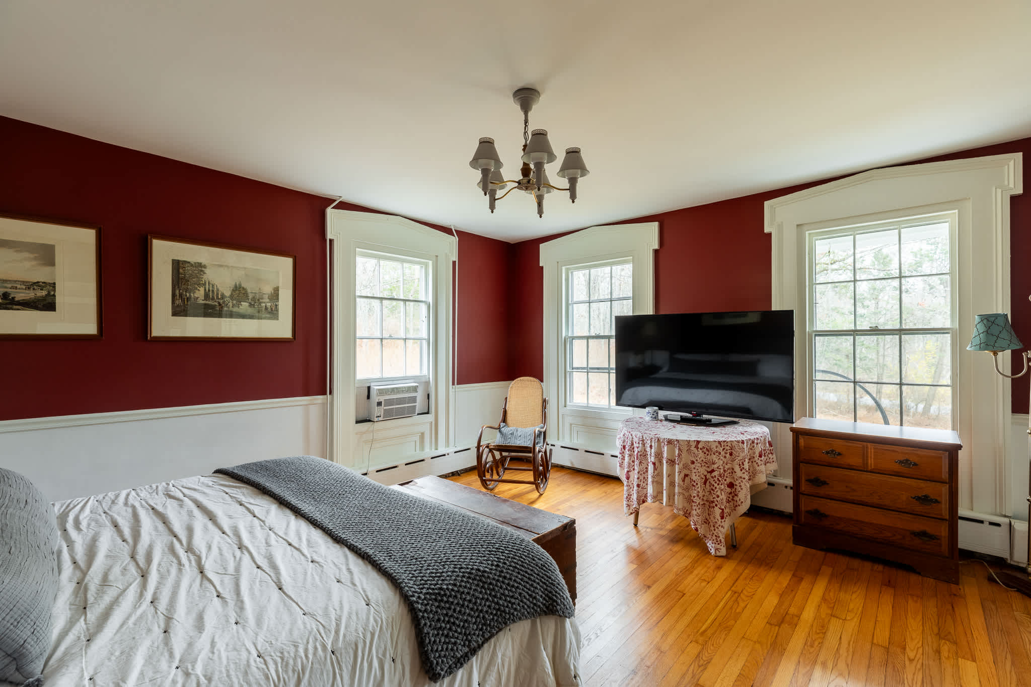 A spacious bedroom with vibrant red walls and white wainscoting, featuring a large bed, multiple windows, and a flat-screen TV on a draped table. A rocking chair and a wooden dresser are also visible, all on polished hardwood floors.