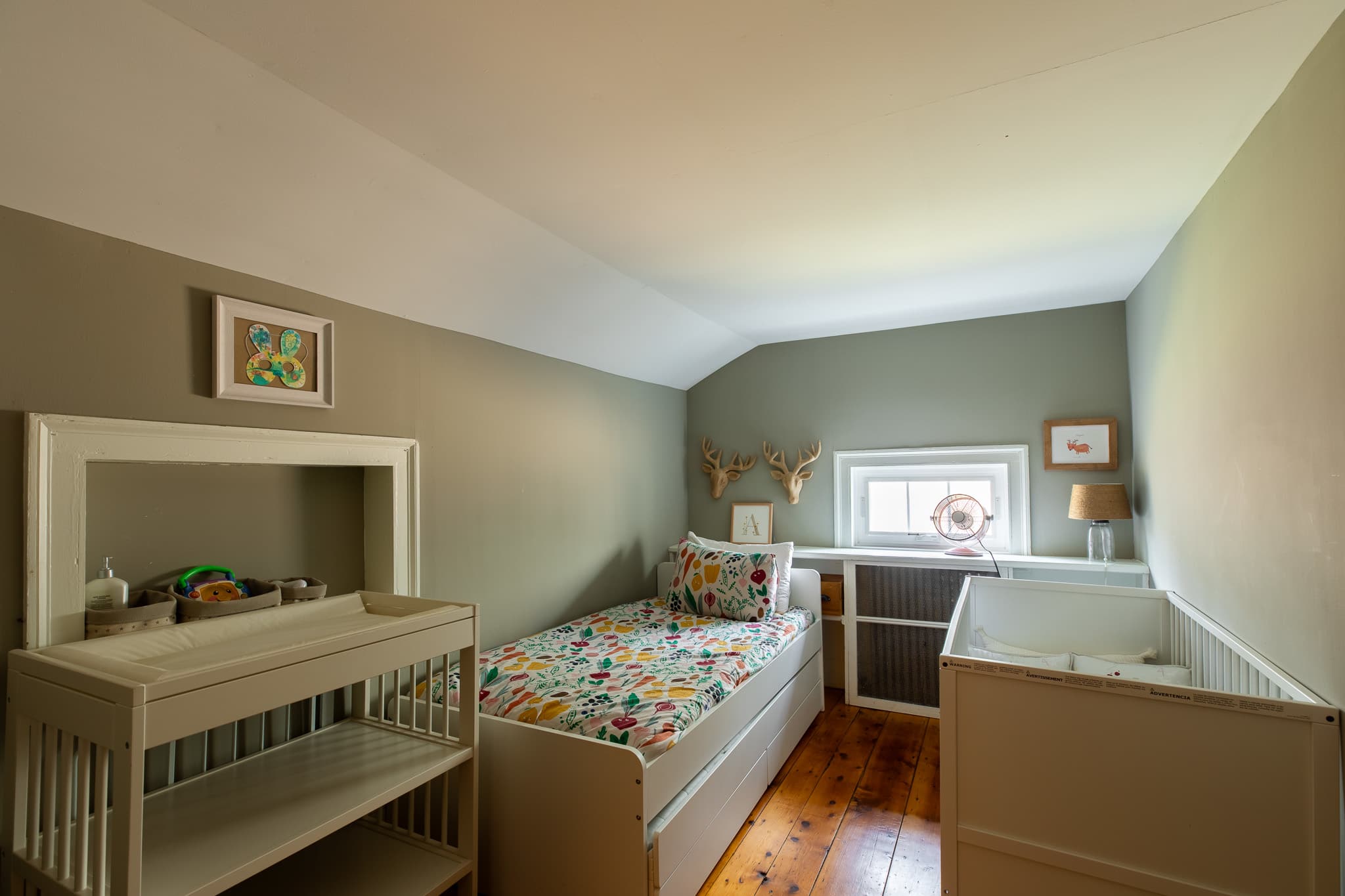 A cozy child's bedroom with light green walls and warm wooden floors, featuring a single bed with colorful floral bedding, a light wood changing table, and a white crib.