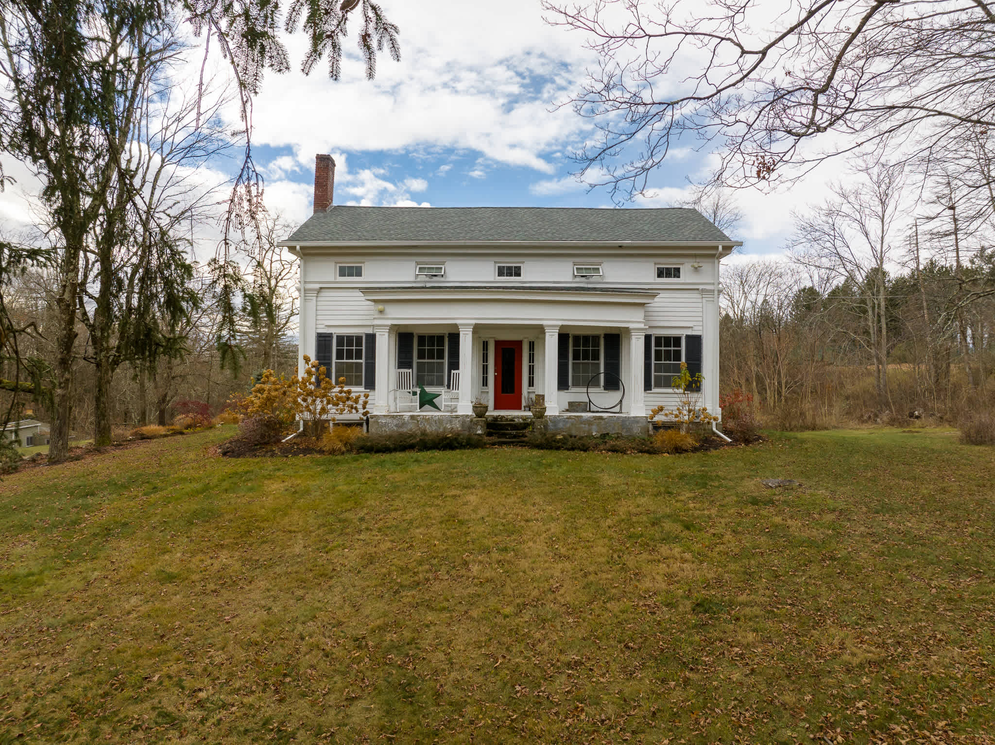 A classic white farmhouse with a dark green roof and a welcoming front porch supported by white columns, viewed from a slight elevation. A vibrant red door stands out between windows adorned with black shutters, surrounded by a grassy lawn and bare trees under a partly cloudy sky.