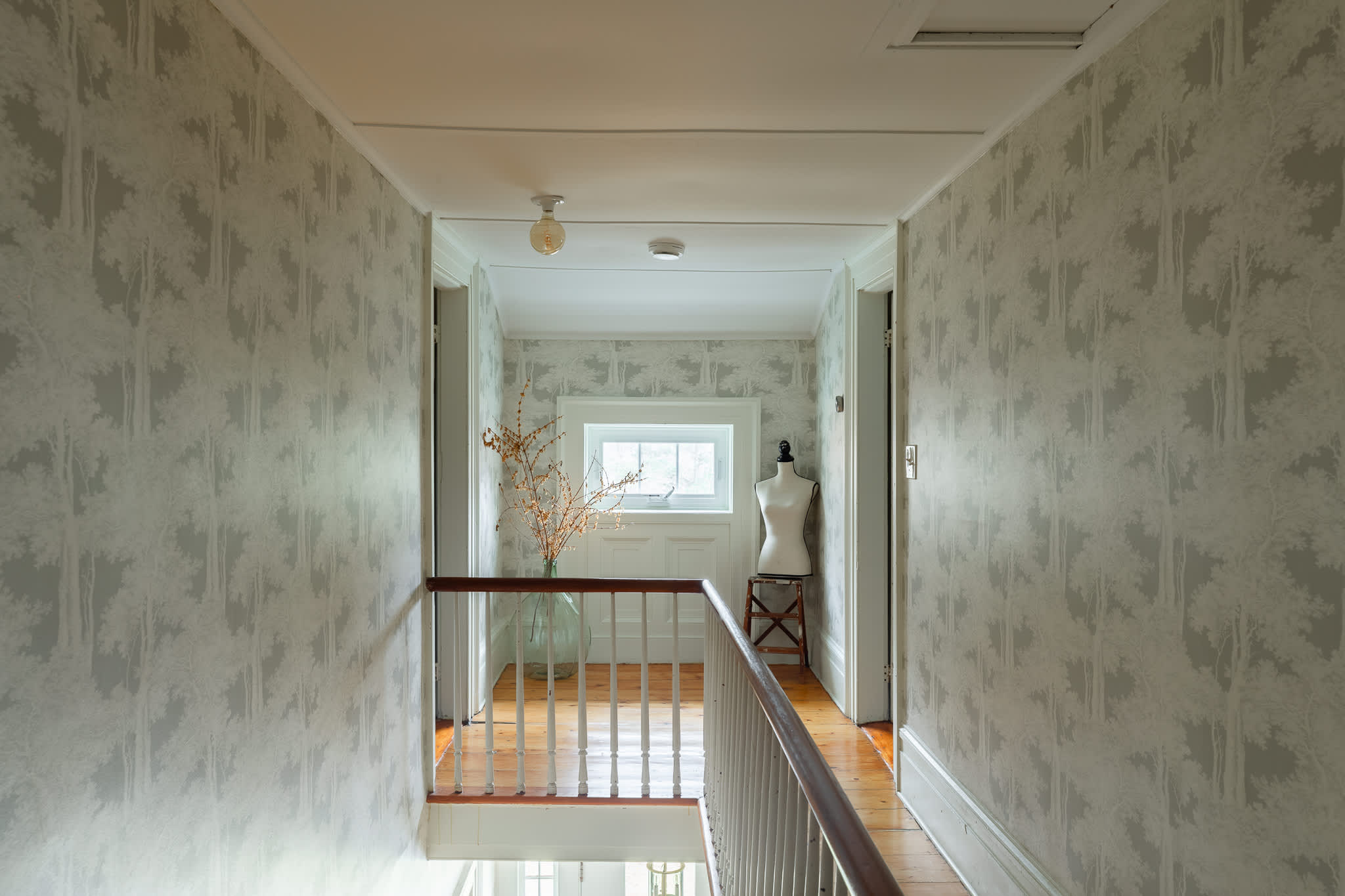 A bright upstairs hallway with light gray tree-patterned wallpaper and warm hardwood floors, featuring a small window and a decorative mannequin. A wooden banister is visible on the right, leading downstairs.