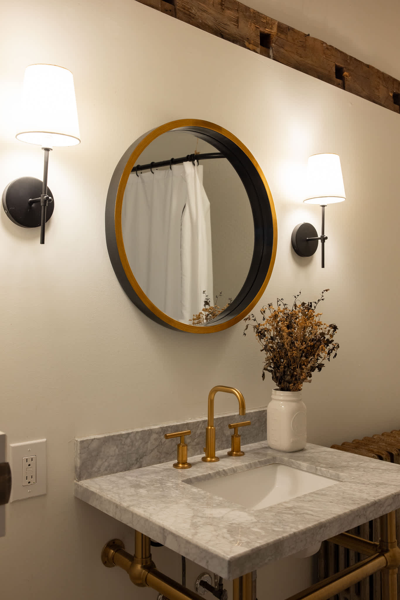A stylish bathroom vanity with a white marble countertop, a gold-framed round mirror, a gold faucet, and two wall sconces, set against a light wall with exposed wooden beams overhead.