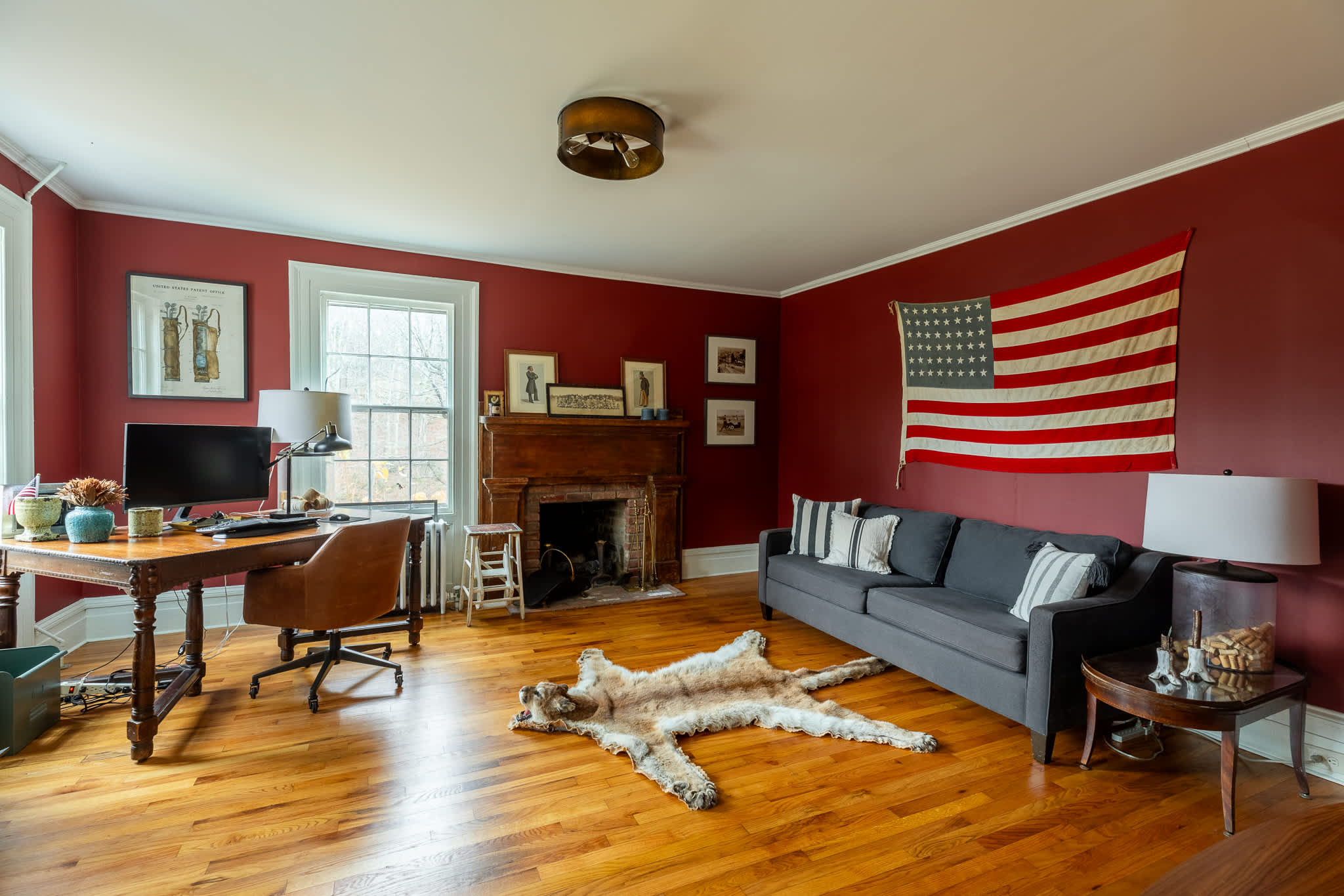 A warm living room with rich red walls and hardwood floors, featuring a gray sofa with striped pillows, a wooden desk with a computer, and a traditional brick fireplace. A large American flag hangs on the wall, and a faux fur rug lies on the floor.