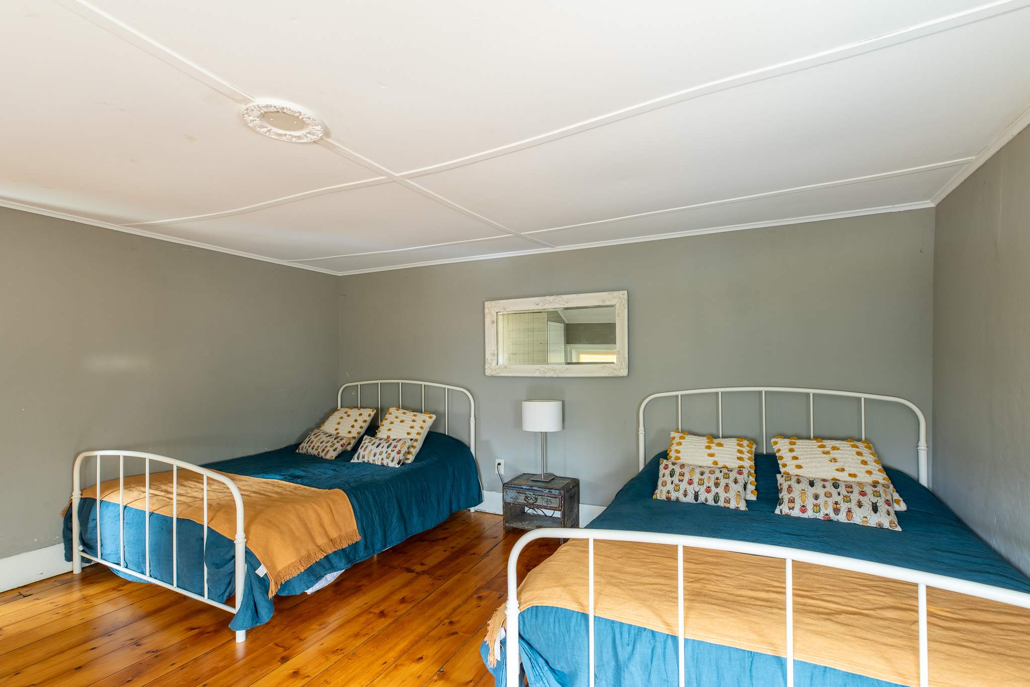 A bright bedroom with light gray walls and polished wooden floors, furnished with two white metal-frame single beds adorned with blue and mustard bedding, and a small white mirror above a rustic nightstand.
