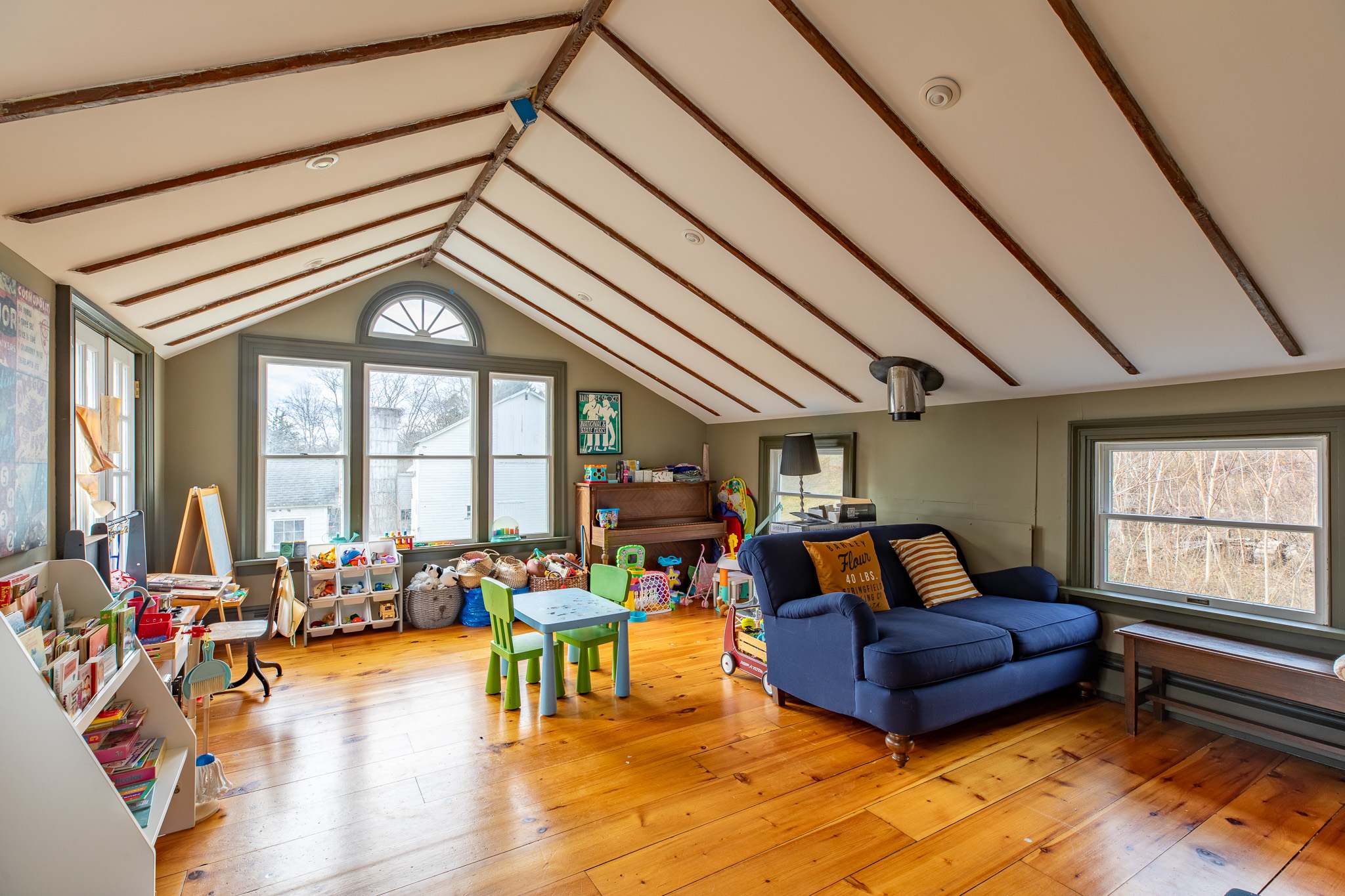 A bright and spacious playroom features an angled ceiling with exposed beams, large windows, a blue sofa, a child-sized table and chairs, and an abundance of toys and books.