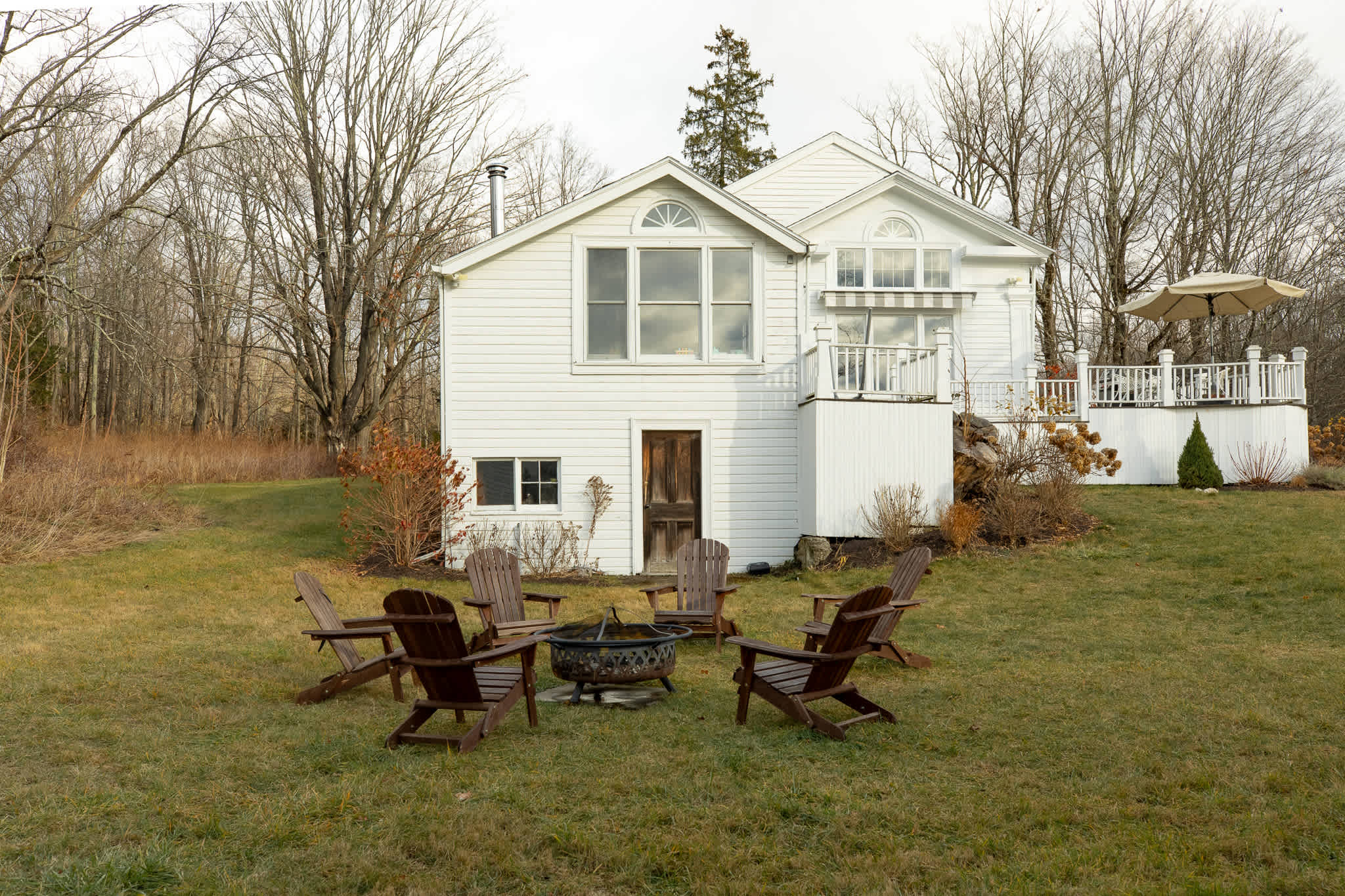 The rear view of a charming white farmhouse with multiple windows and a multi-level deck, overlooking a grassy yard with a fire pit surrounded by dark wooden Adirondack chairs, backed by a forest.