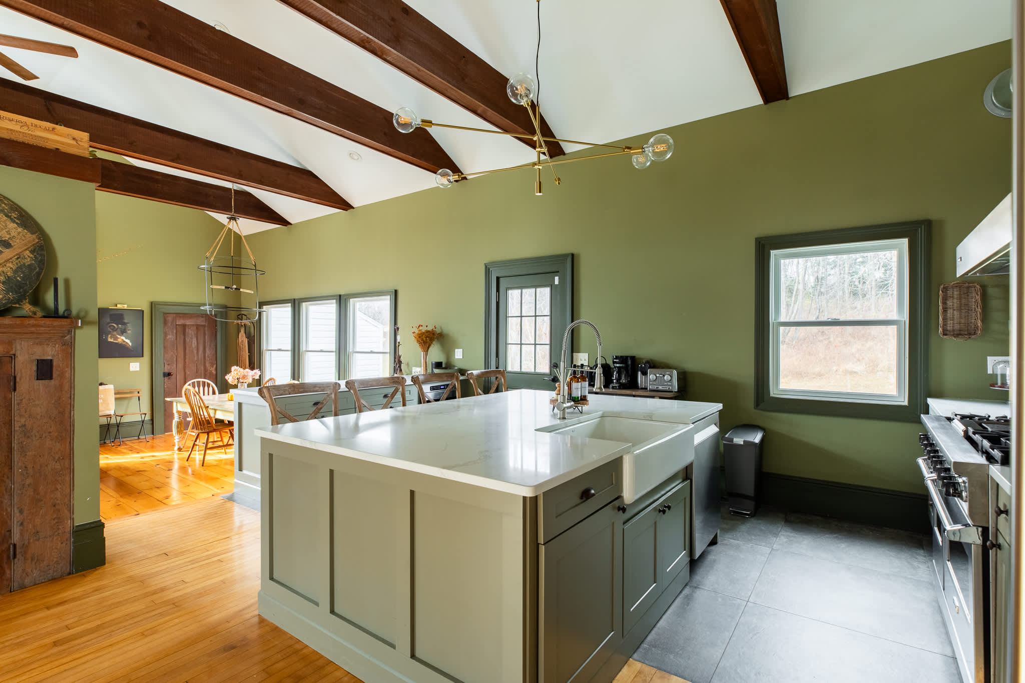 A wide view of the modern kitchen showcases light green cabinets, white countertops, a large island, and stainless steel appliances, with a glimpse of the dining area and its rustic table.