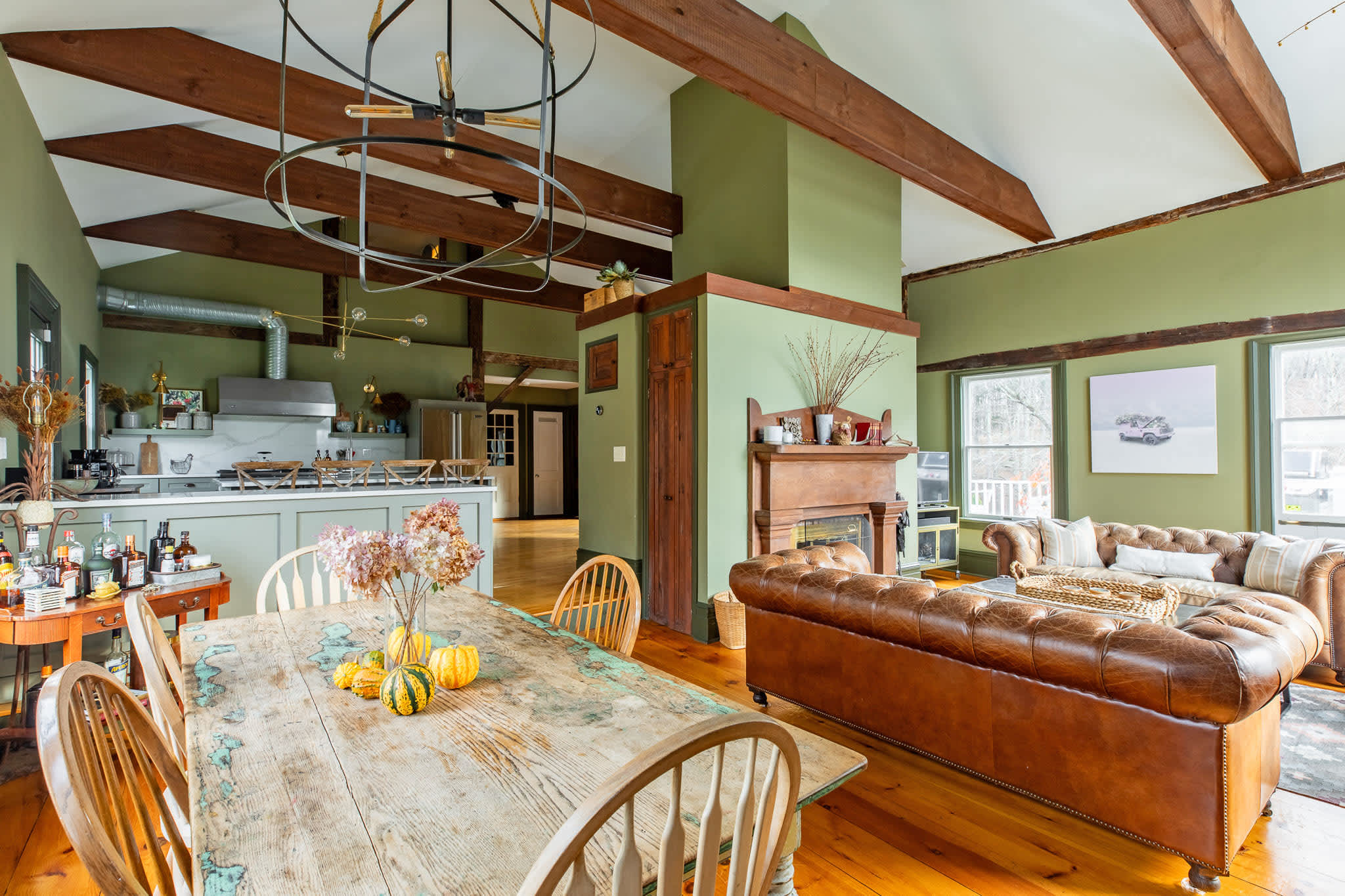 An open-concept living and dining space features olive green walls, exposed wooden beams, a large leather sofa, a rustic dining table, and a view into the modern kitchen.