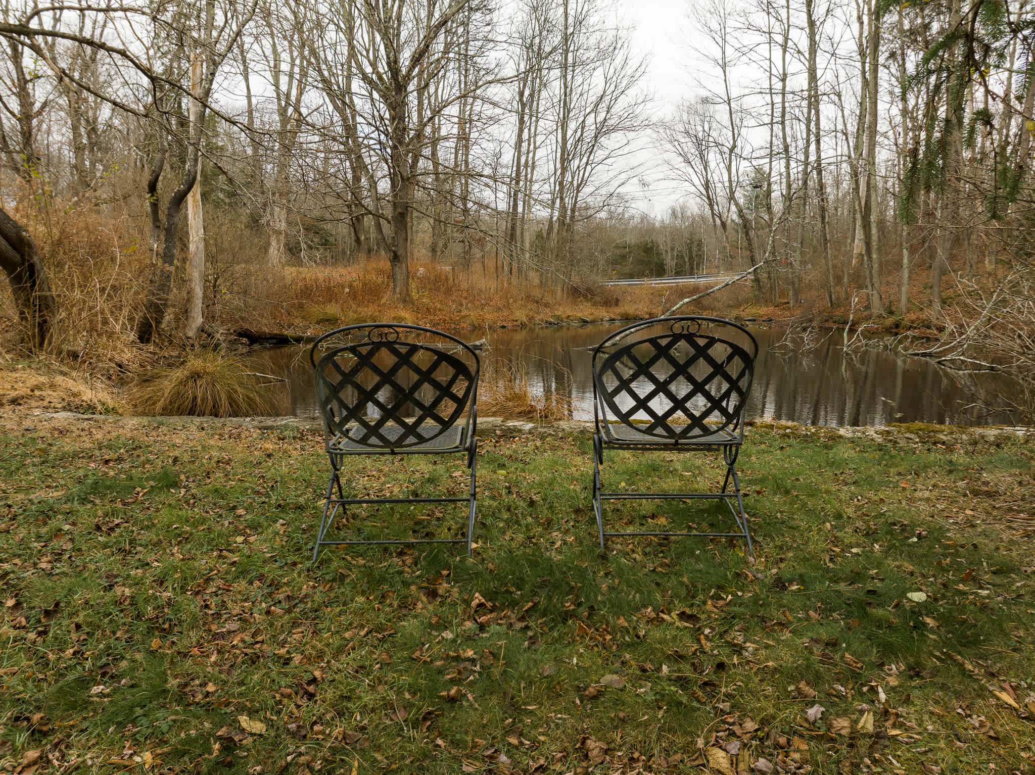A tranquil spot in the yard with two black metal lattice chairs positioned on a grassy bank, offering a peaceful view of a calm, reflective pond or stream bordered by bare trees.