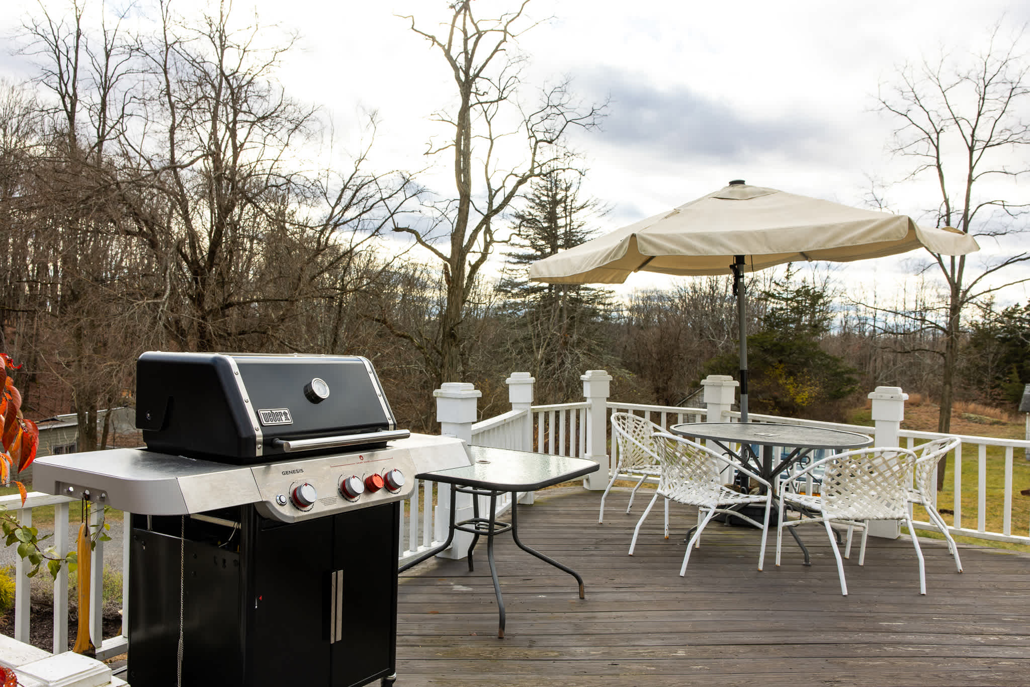 An inviting outdoor wooden deck featuring a black Weber BBQ grill, a round glass-top dining table with white wicker chairs, and a large beige umbrella, surrounded by a white railing.
