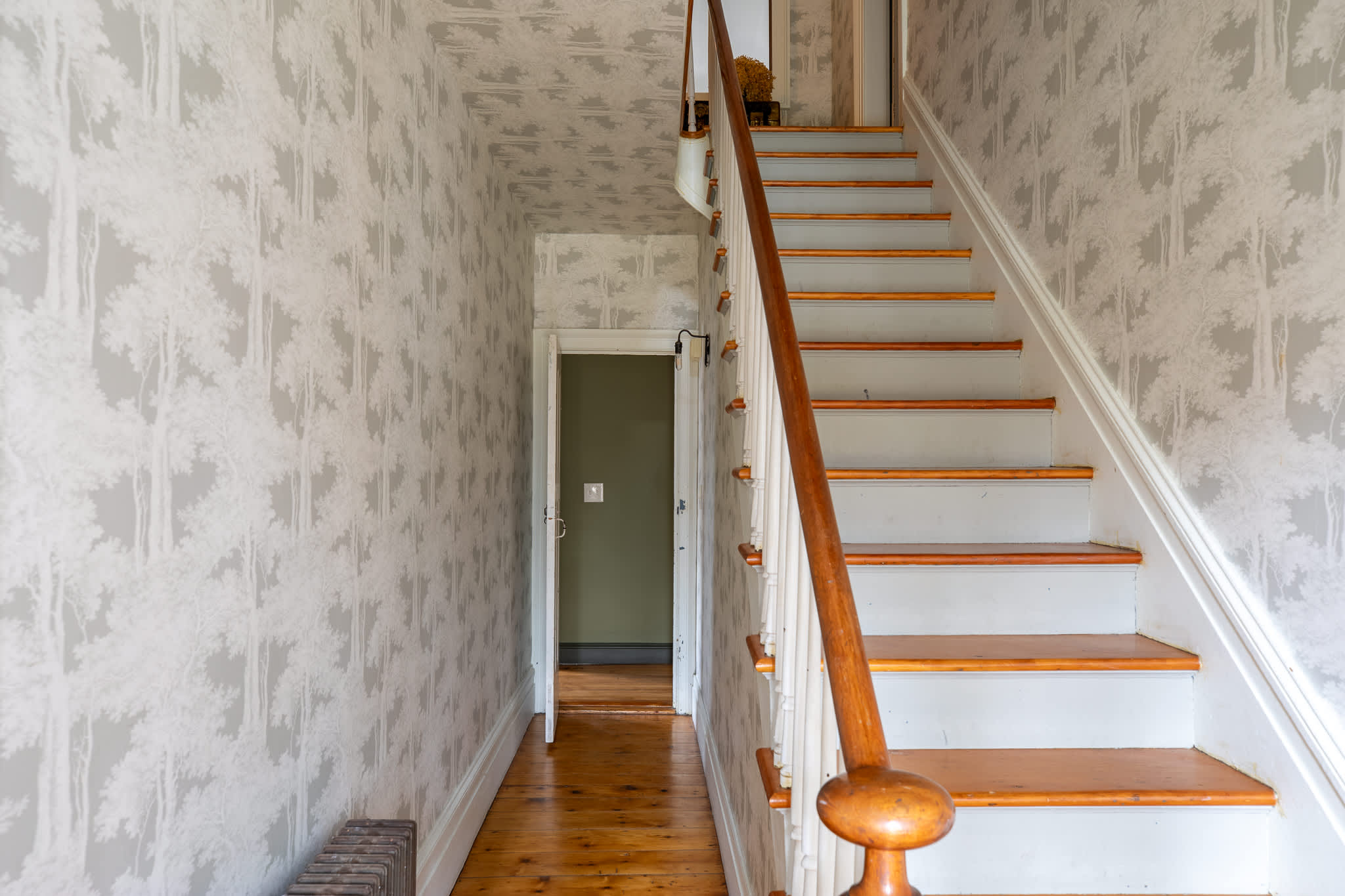A charming hallway and staircase with light gray wallpaper featuring a tree pattern, leading upstairs. The wooden stairs have painted risers and a classic wooden banister, with a doorway visible at the base.