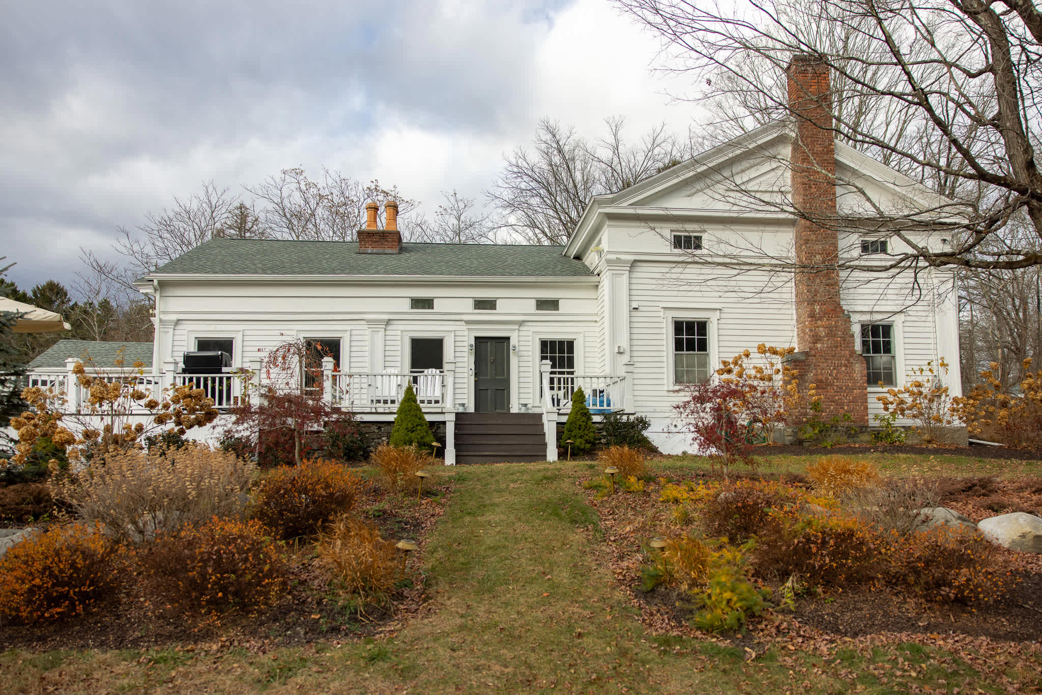 A charming white farmhouse with a dark green roof and a prominent brick chimney, viewed from a corner angle. A wooden deck with white railings extends from the left, and a path leads to dark gray steps at the main entrance, surrounded by autumn-colored shrubs and a grassy lawn.