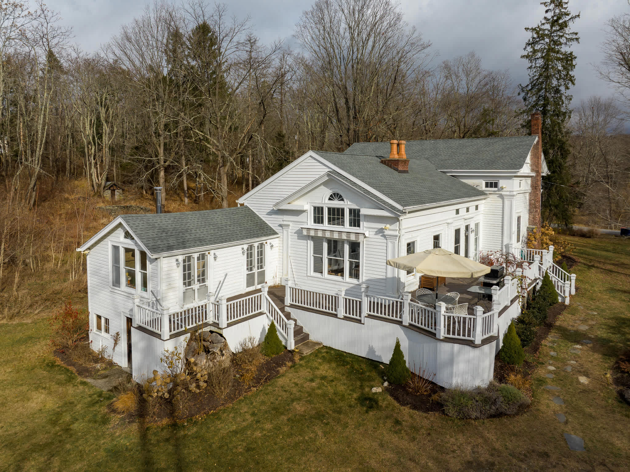 An expansive aerial view of a large white farmhouse with a gray roof and numerous windows, showcasing its complex structure, surrounding bare trees, and extensive wooden decks with outdoor furniture.