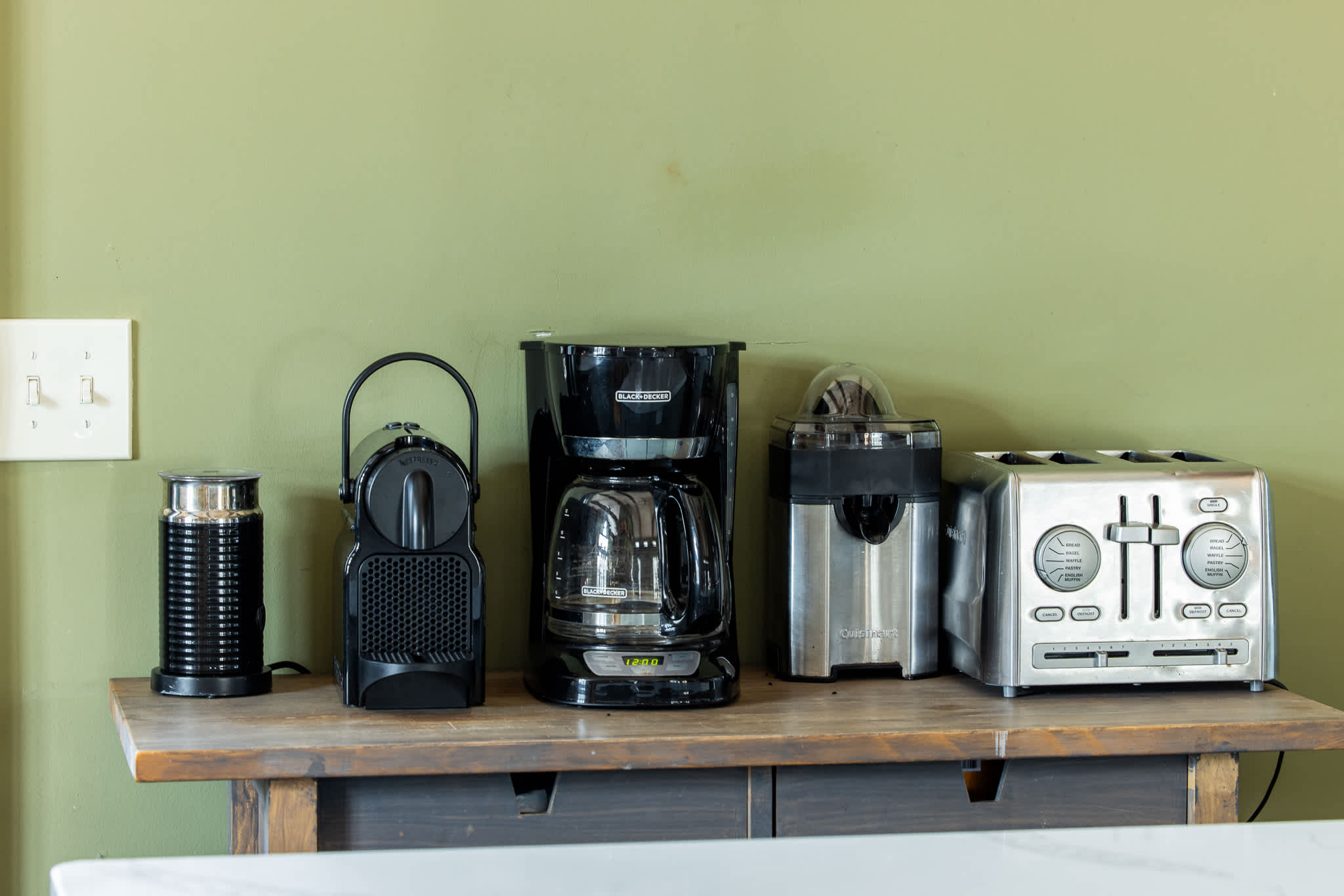 A dedicated coffee station on a wooden counter features a Nespresso machine, a drip coffee maker, a citrus juicer, and a toaster, set against an olive green wall.