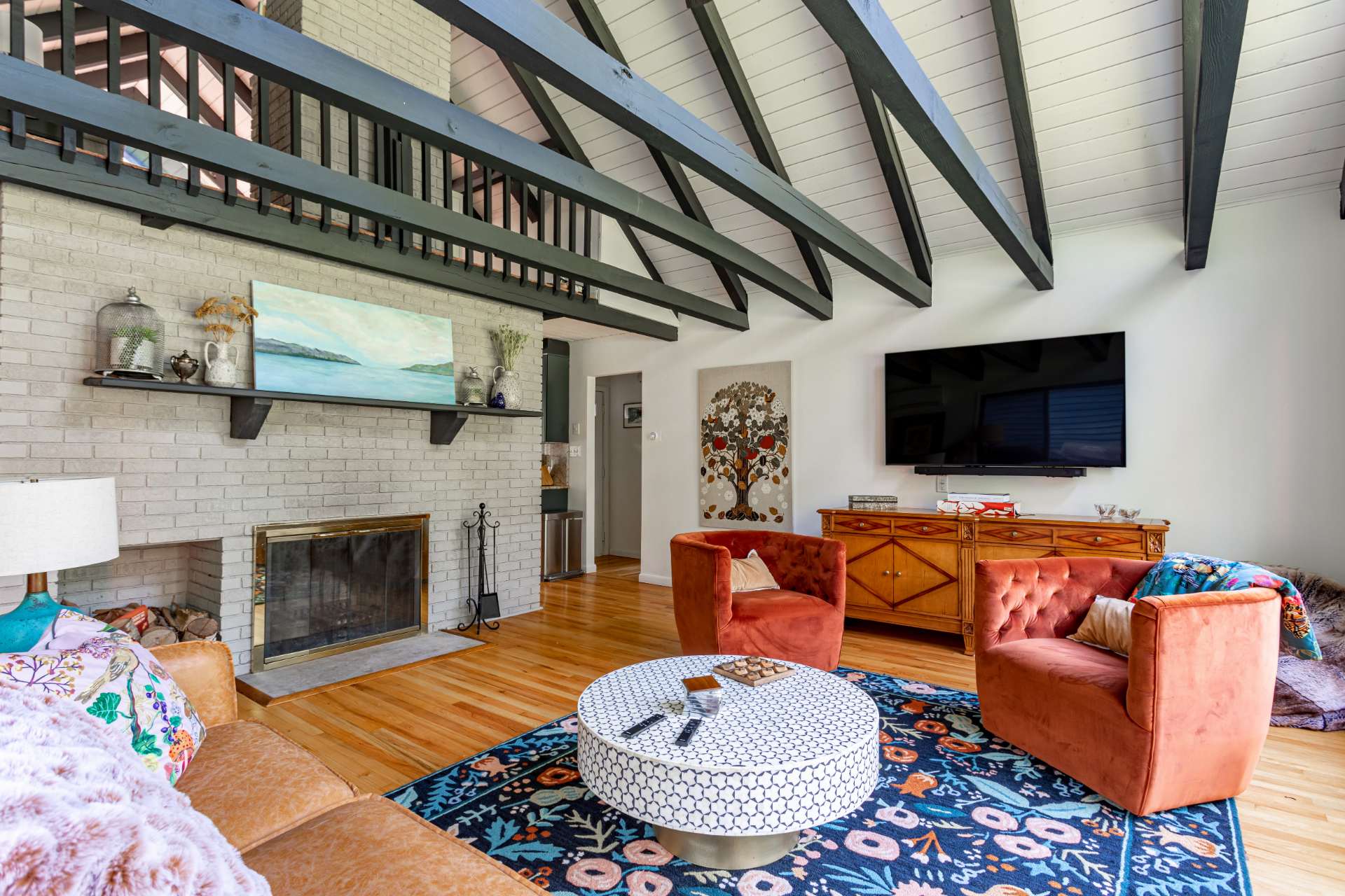 Cozy living room featuring a white brick fireplace with a wooden mantel, a large wall-mounted TV, and a dark blue patterned rug. Exposed dark beams contrast with the white vaulted ceiling, and a balcony overlooks the space.
