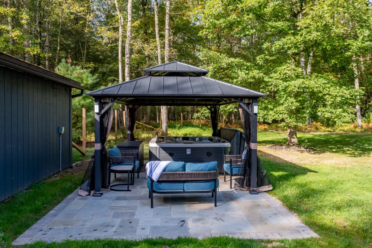 An outdoor hot tub is situated under a dark metal gazebo with sheer black curtains, surrounded by blue cushioned patio furniture on a stone paver patio. Lush green grass and a dense forest line the background.