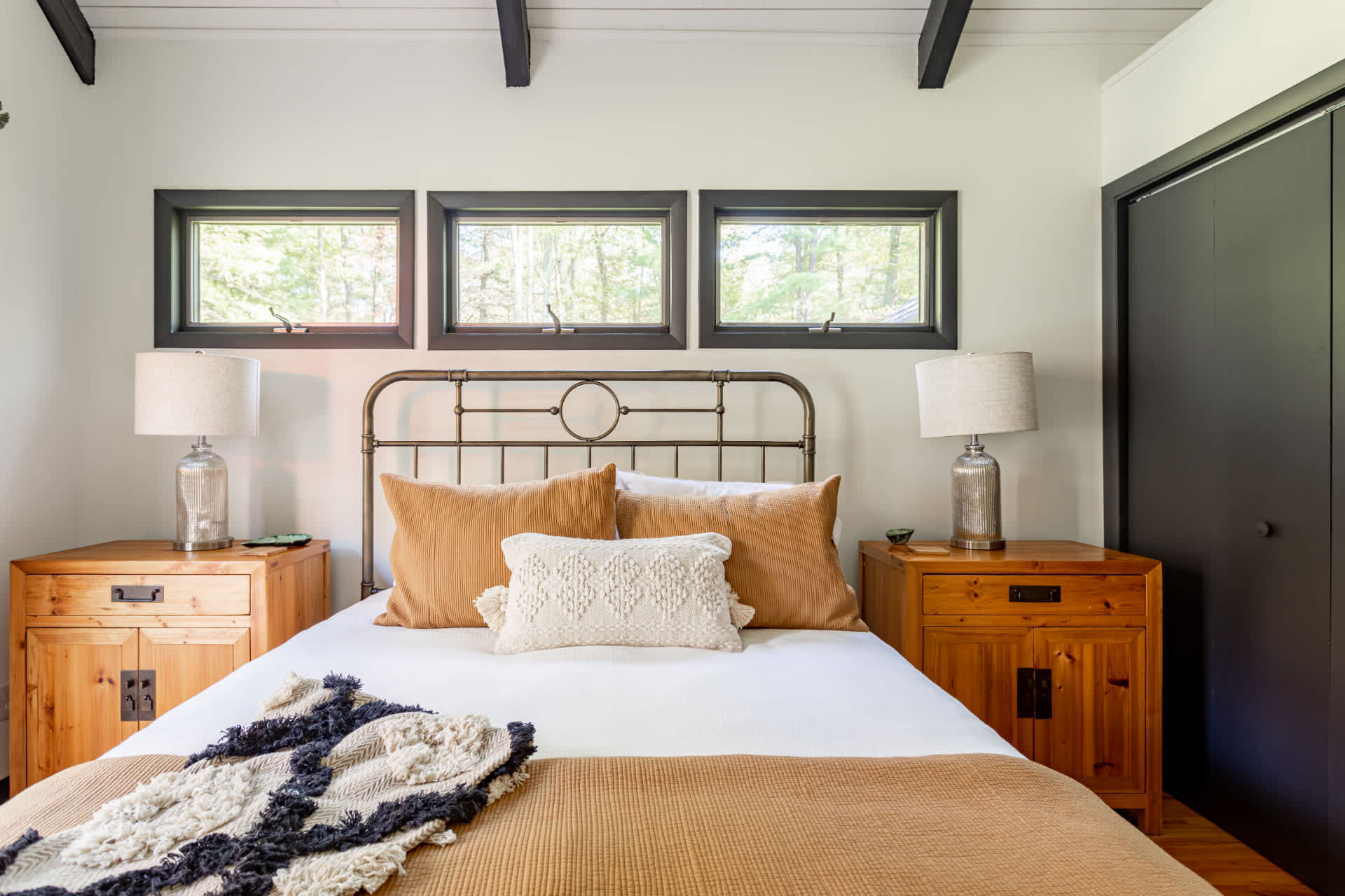 Master bedroom featuring a metal bed frame with white and rust-colored bedding, flanked by wooden nightstands with lamps. Three horizontal windows are positioned above the bed, and the ceiling has dark exposed beams.
