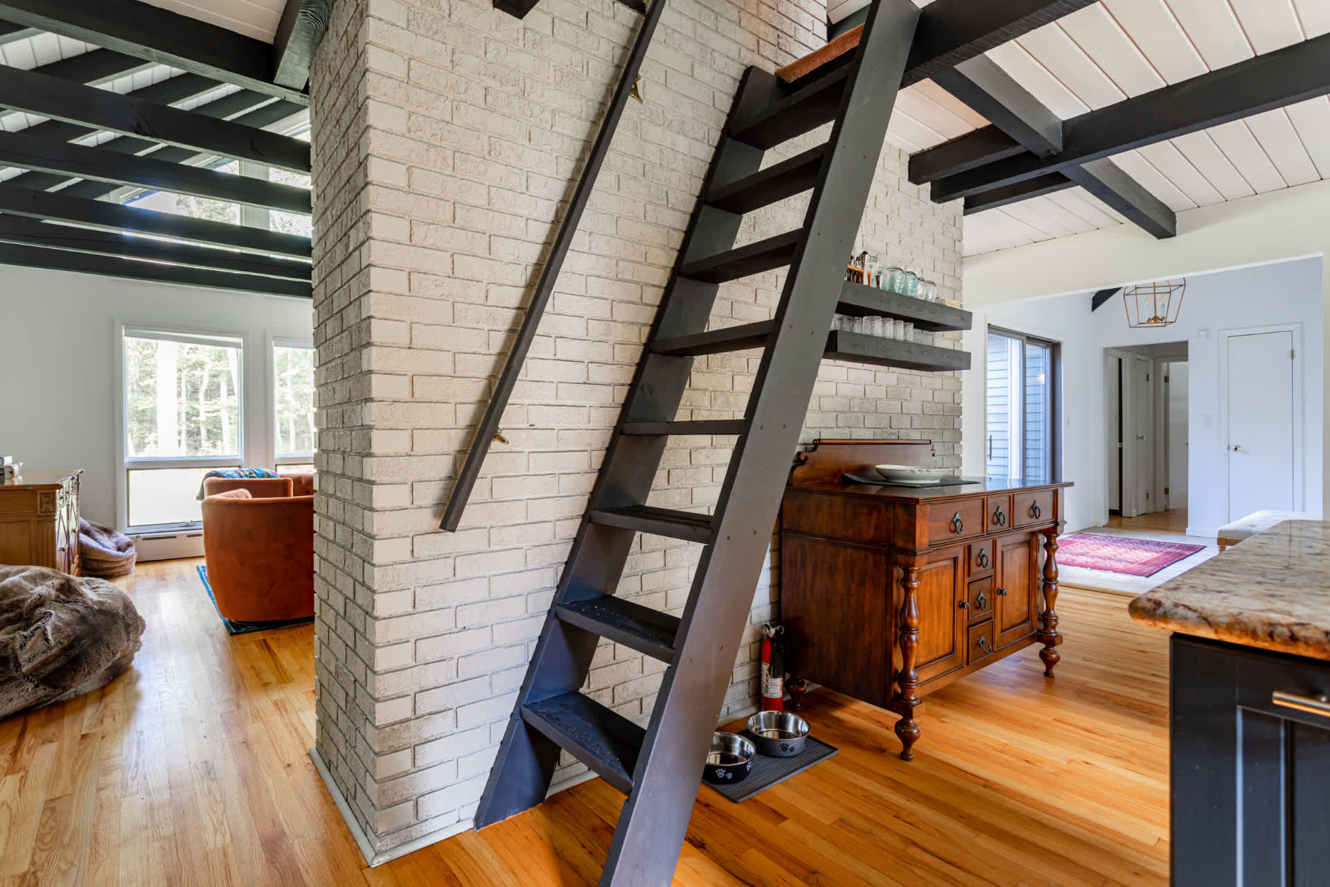 Interior view showing a black wooden ladder-style staircase leading to an upper level, adjacent to a light-colored brick column. Hardwood floors are visible, and a wooden cabinet is beneath the stairs.