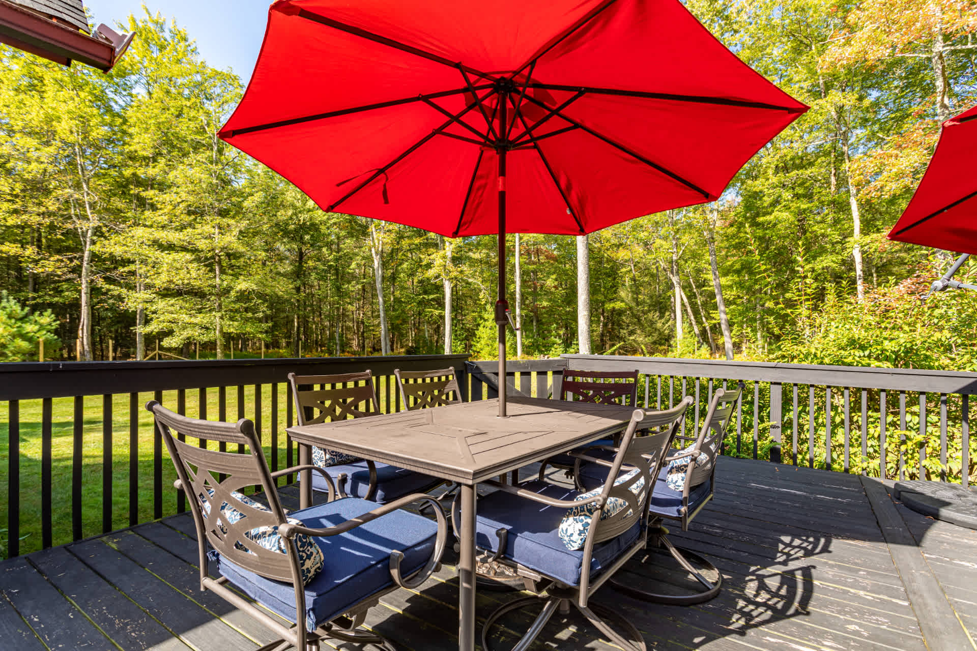 A dark wooden deck features a metal outdoor dining set with six chairs, each with blue cushions and patterned pillows, shaded by a large red umbrella. A lush green forest provides a natural backdrop.