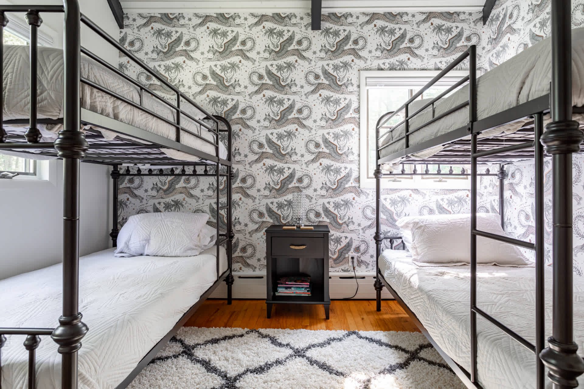 Kids' bedroom with two black metal bunk beds, a dark nightstand between them, and patterned wallpaper featuring animals. A shaggy area rug covers part of the hardwood floor.