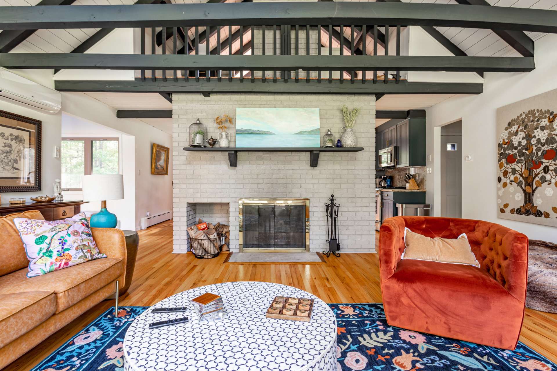 Cozy living room centered around a white brick fireplace with a dark mantel and a painting above, flanked by a brown leather sofa and an orange armchair, with a loft area visible overhead.