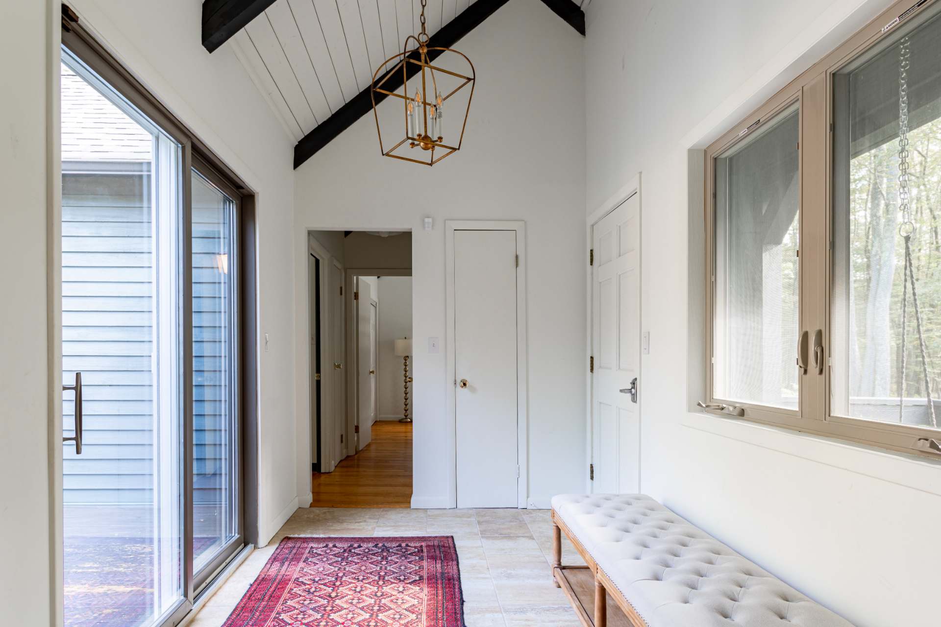 Bright entryway with a high vaulted ceiling, exposed dark beams, a gold chandelier, a long upholstered bench, and a red patterned rug on tiled floors, leading to other rooms.
