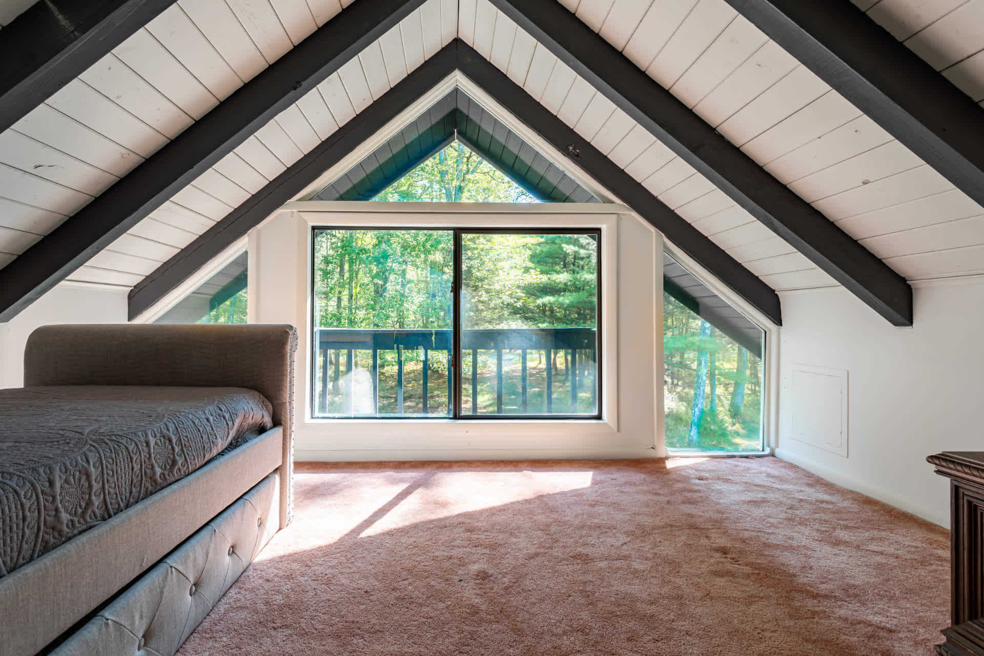 Loft area with a daybed and trundle, light brown carpet, and a large A-frame window offering views of the outdoors. The ceiling has dark exposed beams.